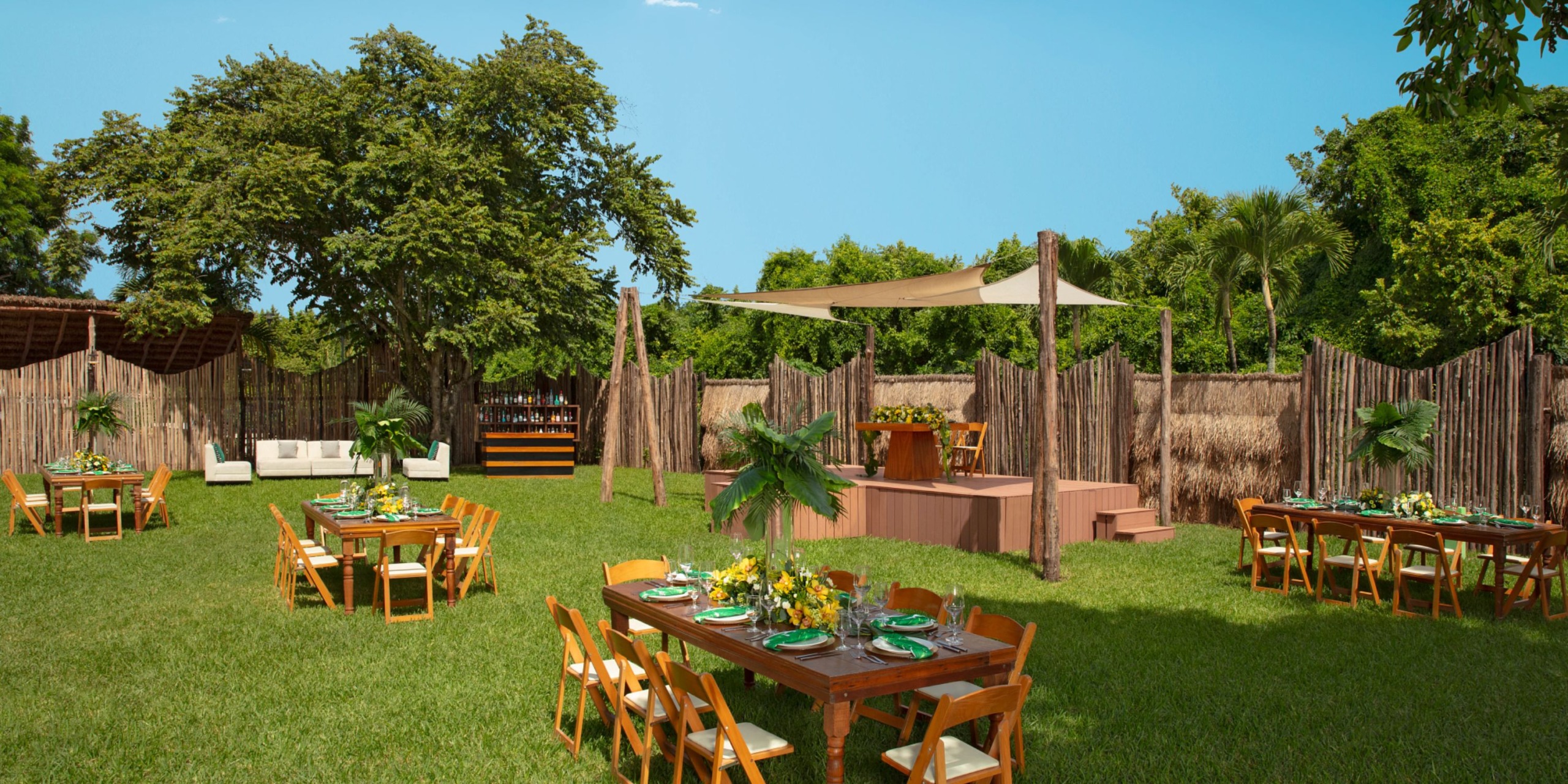 An event setup featuring the Dreams Cozumel Cape collection atop a grassy area, showcasing several wooden tables and chairs. The tables are adorned with floral centerpieces. In the background, there's a stage covered by a canopy and accompanied by a bar, all nestled among trees.