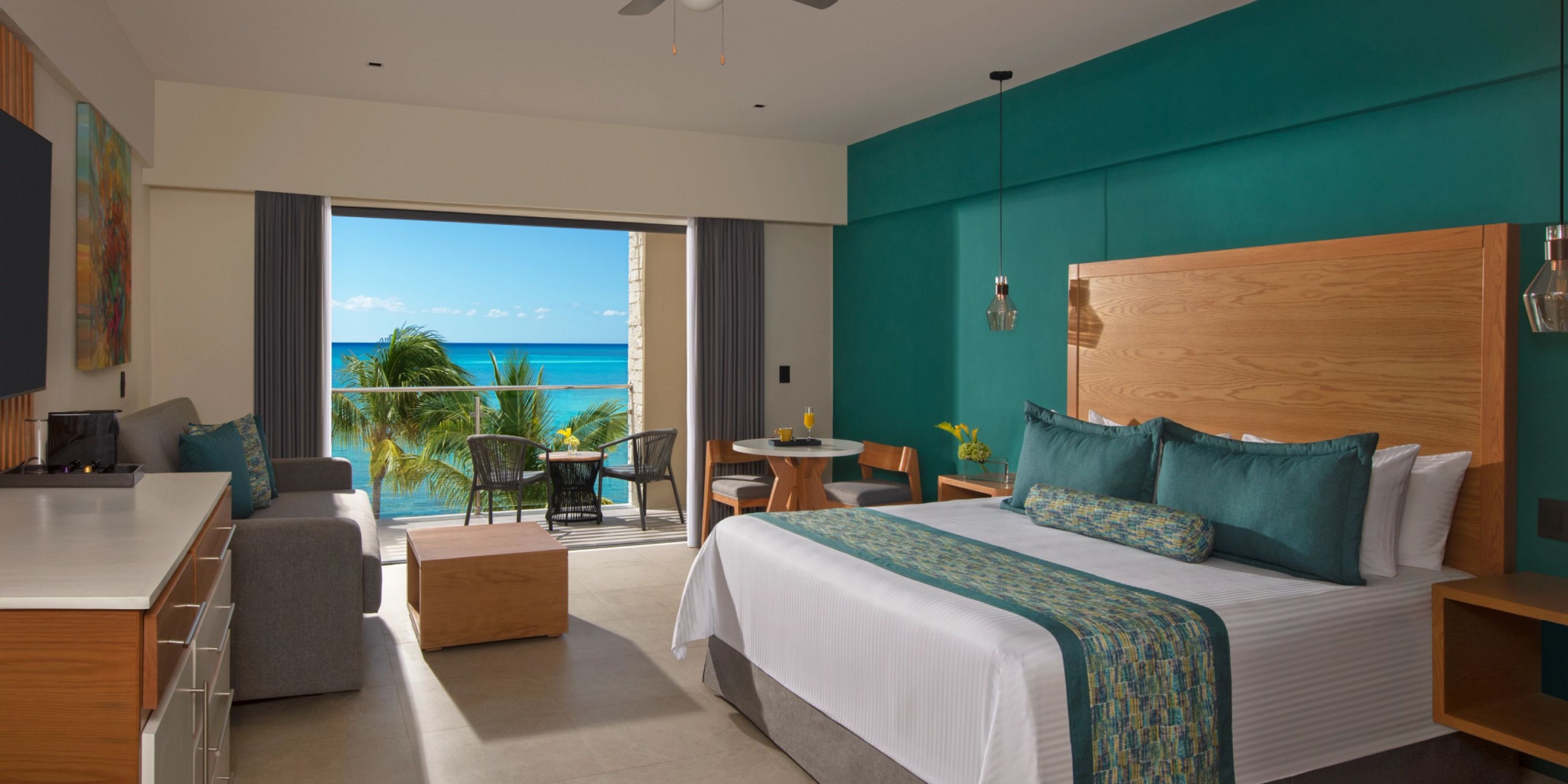 Experience the Dreams Cozumel Cape, featuring a spacious room with a large bed, sofa, and small table. Balcony doors reveal stunning views of palm trees and the ocean. The room is adorned with bright teal accents and modern decor for a perfect retreat.