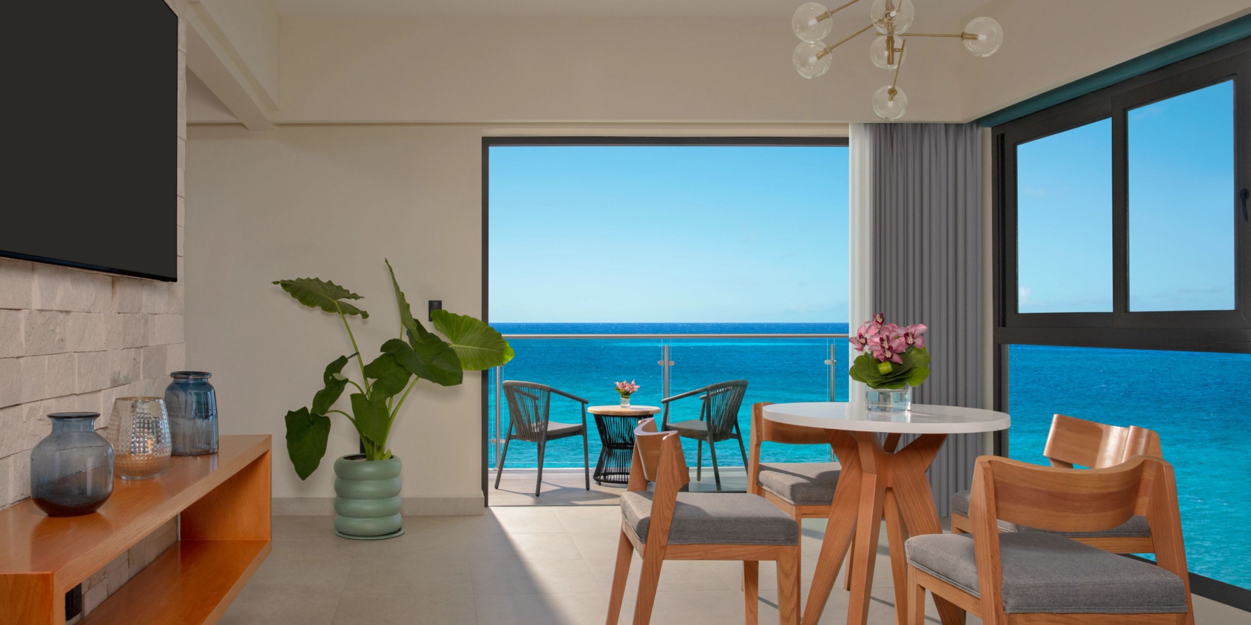 Dreams Cozumel Cape living room featuring a round table, wooden chairs, potted plants, and open balcony doors providing a beautiful ocean view.