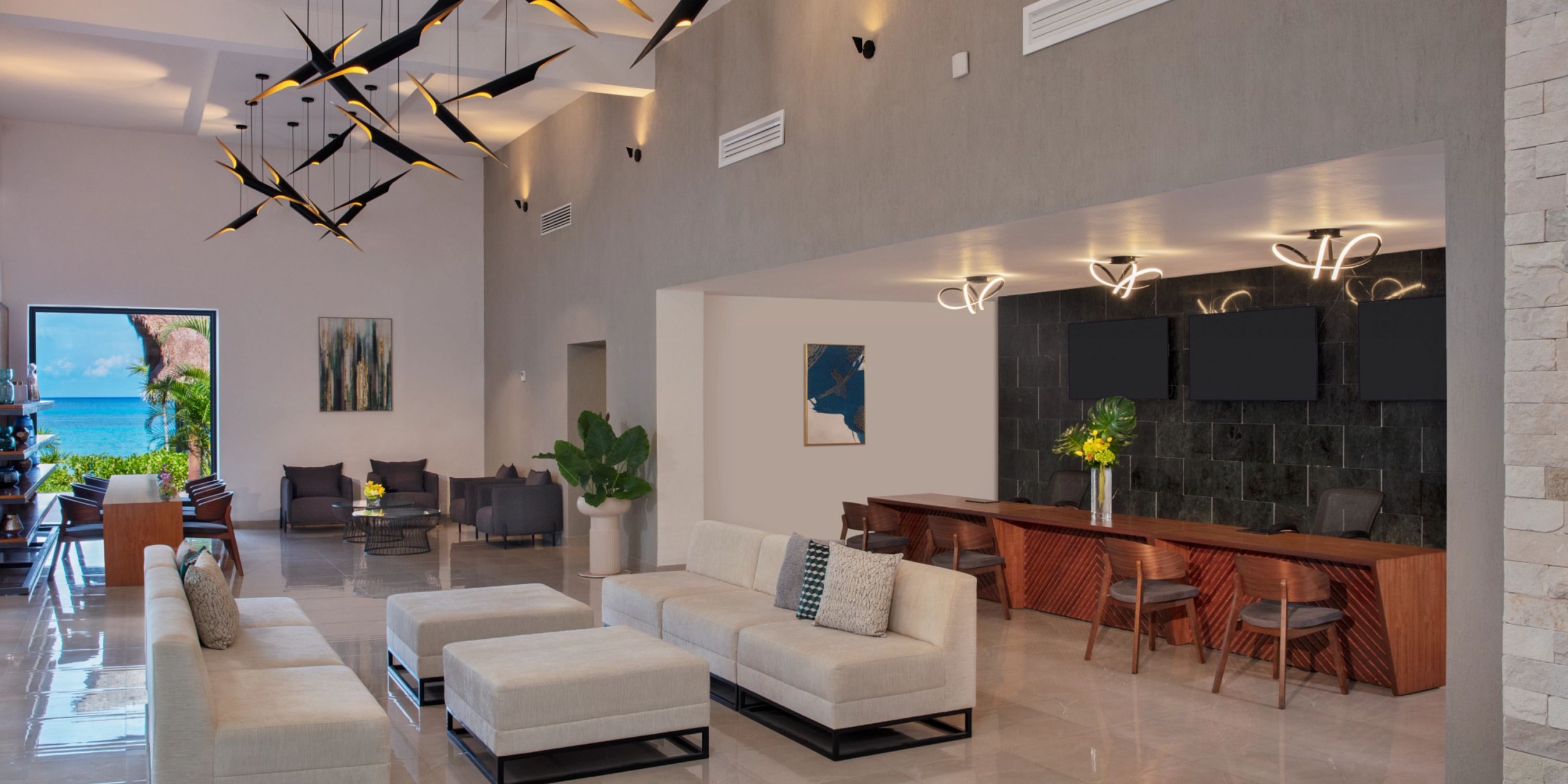 The Dreams Cozumel Cape boasts a contemporary lobby featuring beige sofas, a reception desk, and a black stone wall. Decorative ceiling lights enhance the ambiance, while the ocean view can be admired through the entrance.