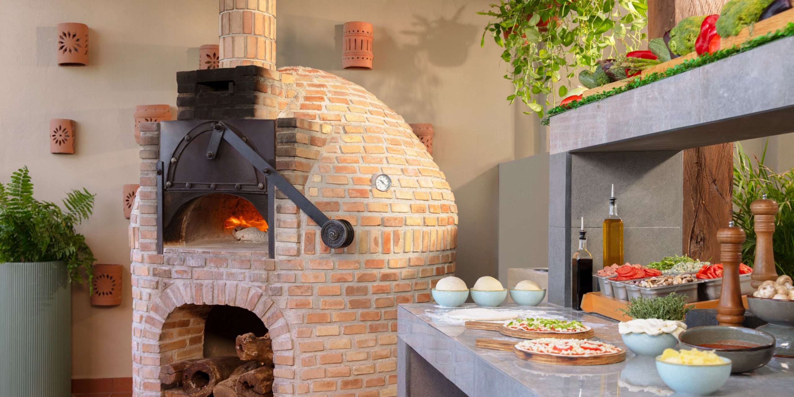 A Dreams Cozumel Cape brick pizza oven beautifully accented by a fire and surrounded by lush plants. Close by, a counter is topped with all the essentials for pizza-making, including dough, tomatoes, and cheese.