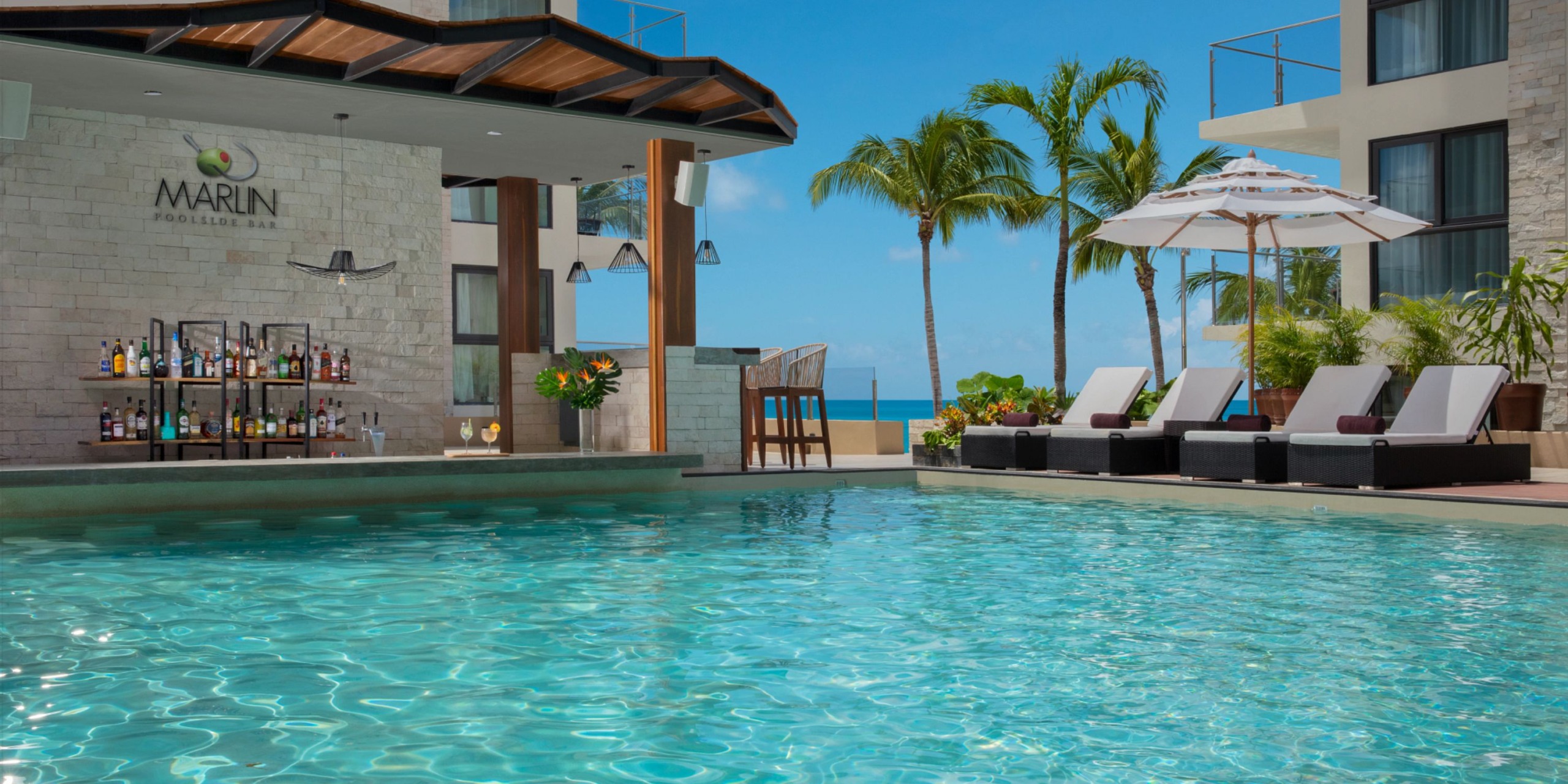 The Dreams Cozumel Cape offers an outdoor pool area complete with lounge chairs, a poolside bar, and ocean views, surrounded by palm trees under a clear blue sky.