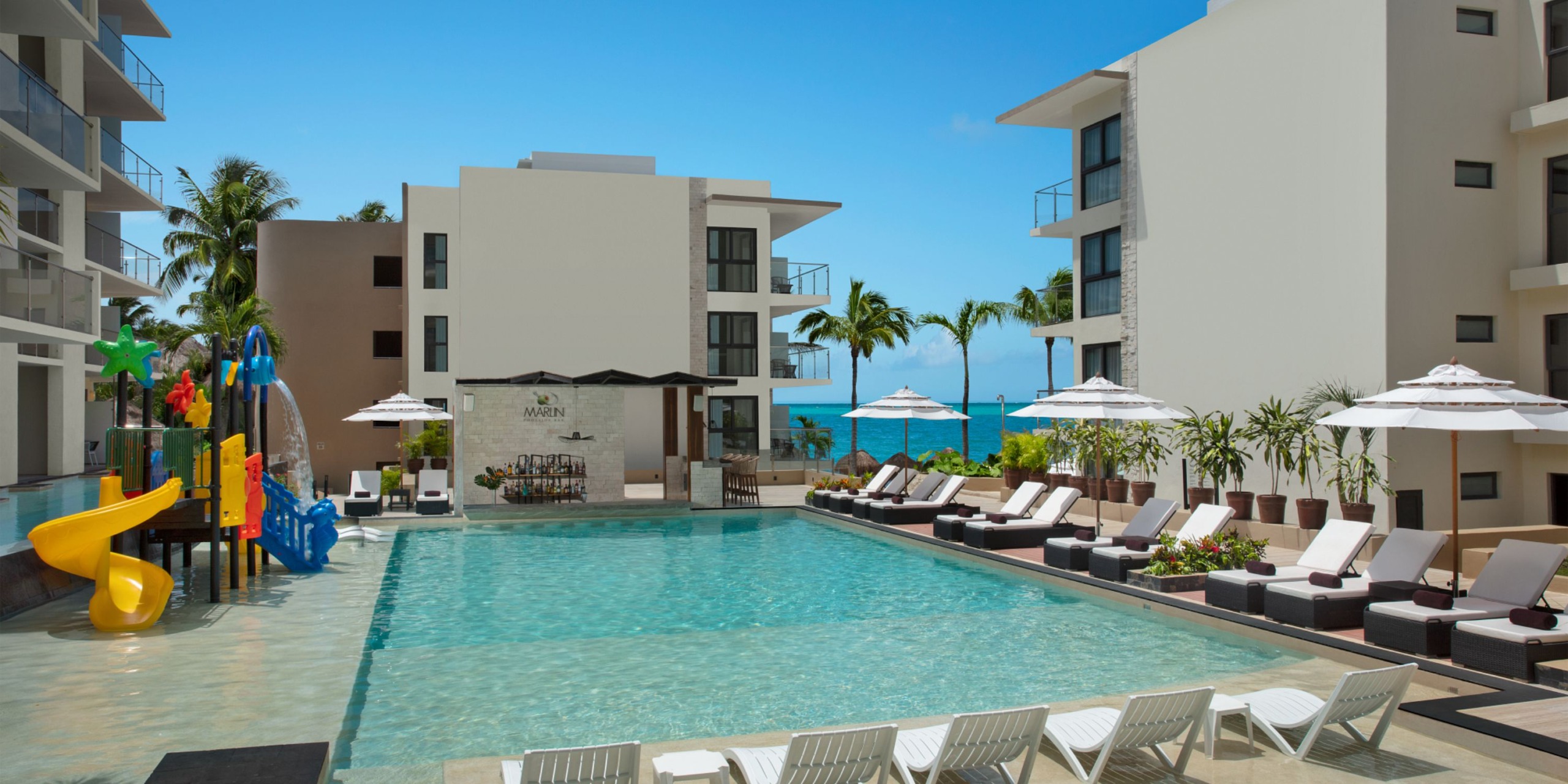 The Dreams Cozumel Cape features a hotel pool area complete with lounge chairs, umbrellas, and a water play structure. White buildings and palm trees surround the area, offering views of the ocean against a backdrop of clear skies.