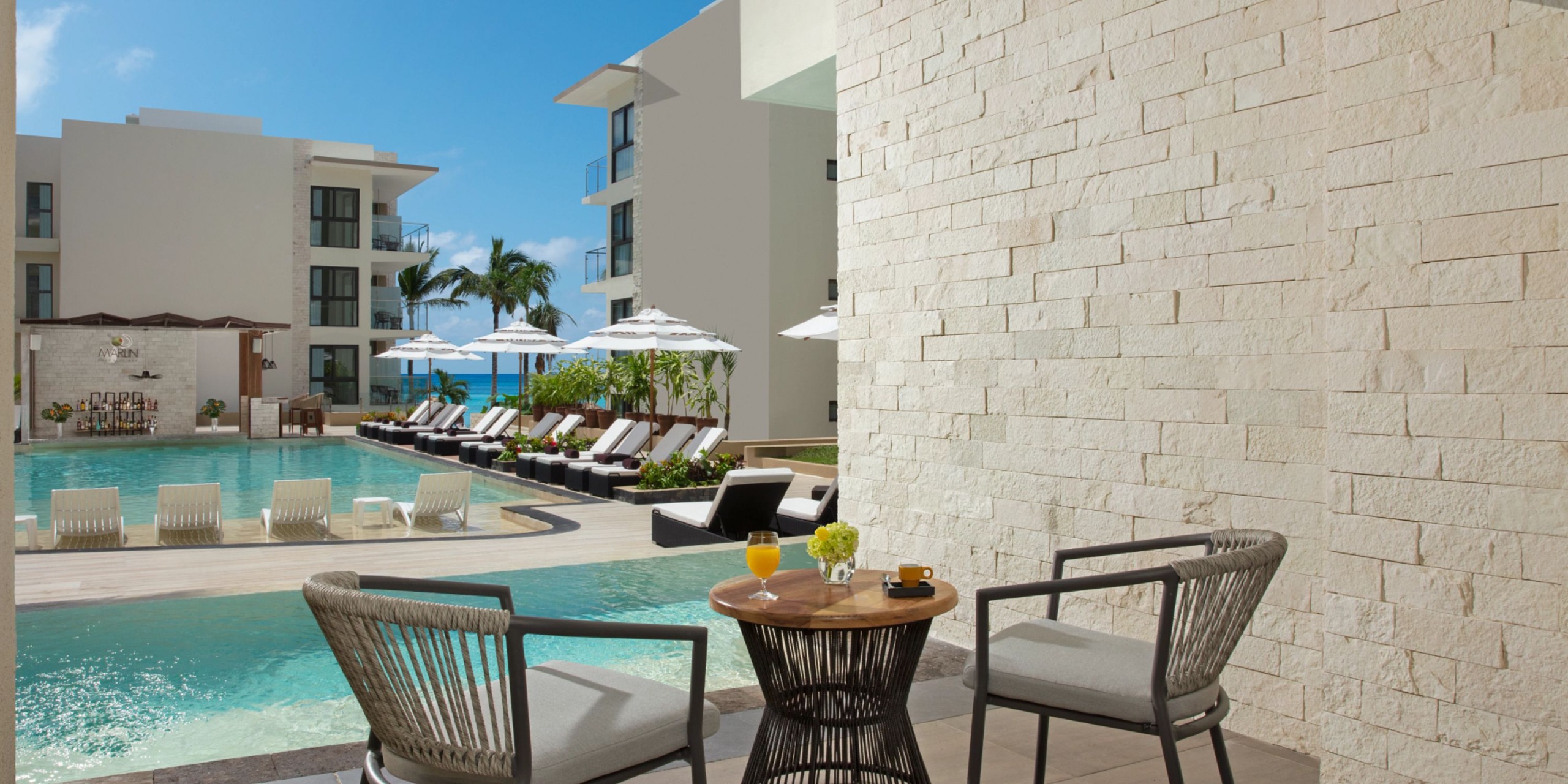 Two chairs and a table with drinks on the poolside patio at Dreams Cozumel Cape overlook a swimming pool and sun loungers, with modern white buildings and palm trees providing a stunning backdrop.