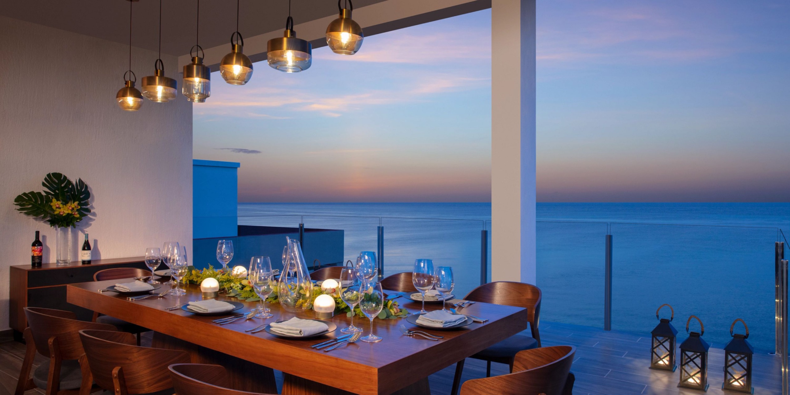 Elegant Dreams Cozumel Cape dining table set for eight on a terrace overlooking the ocean at sunset, featuring modern pendant lights and decorative lanterns.
