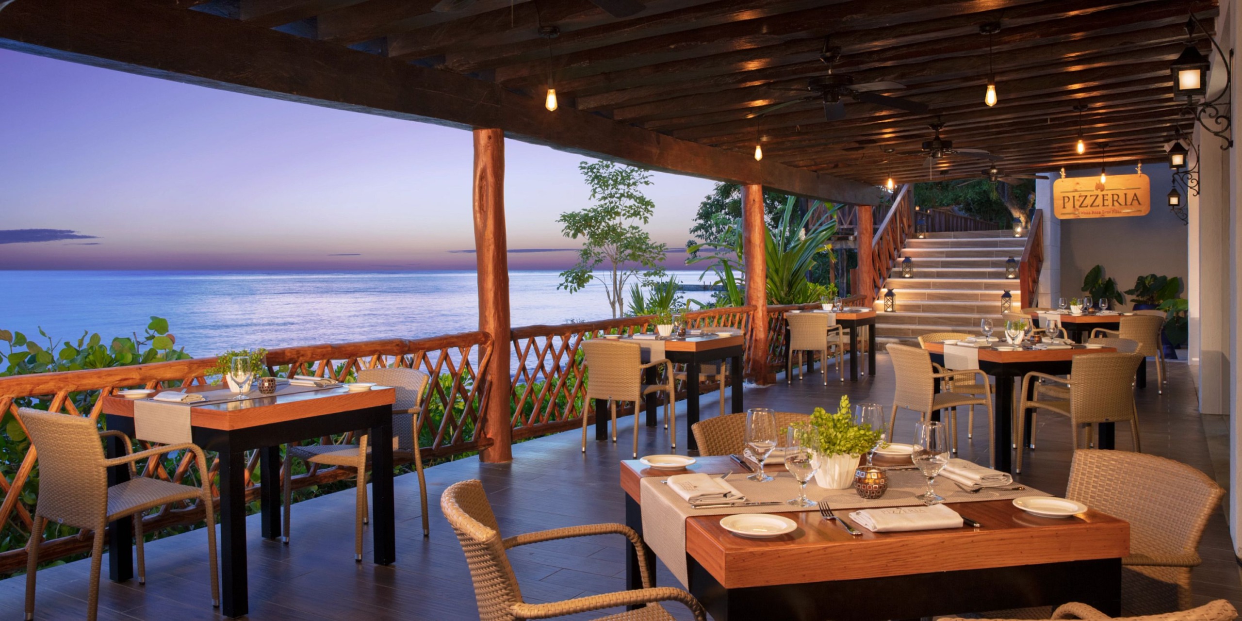 The Dreams Cozumel Cape offers an outdoor restaurant patio with tables set for dining, featuring a breathtaking ocean view in the background and a staircase leading upwards. The atmosphere is serene, highlighted by a tranquil dusk sky.