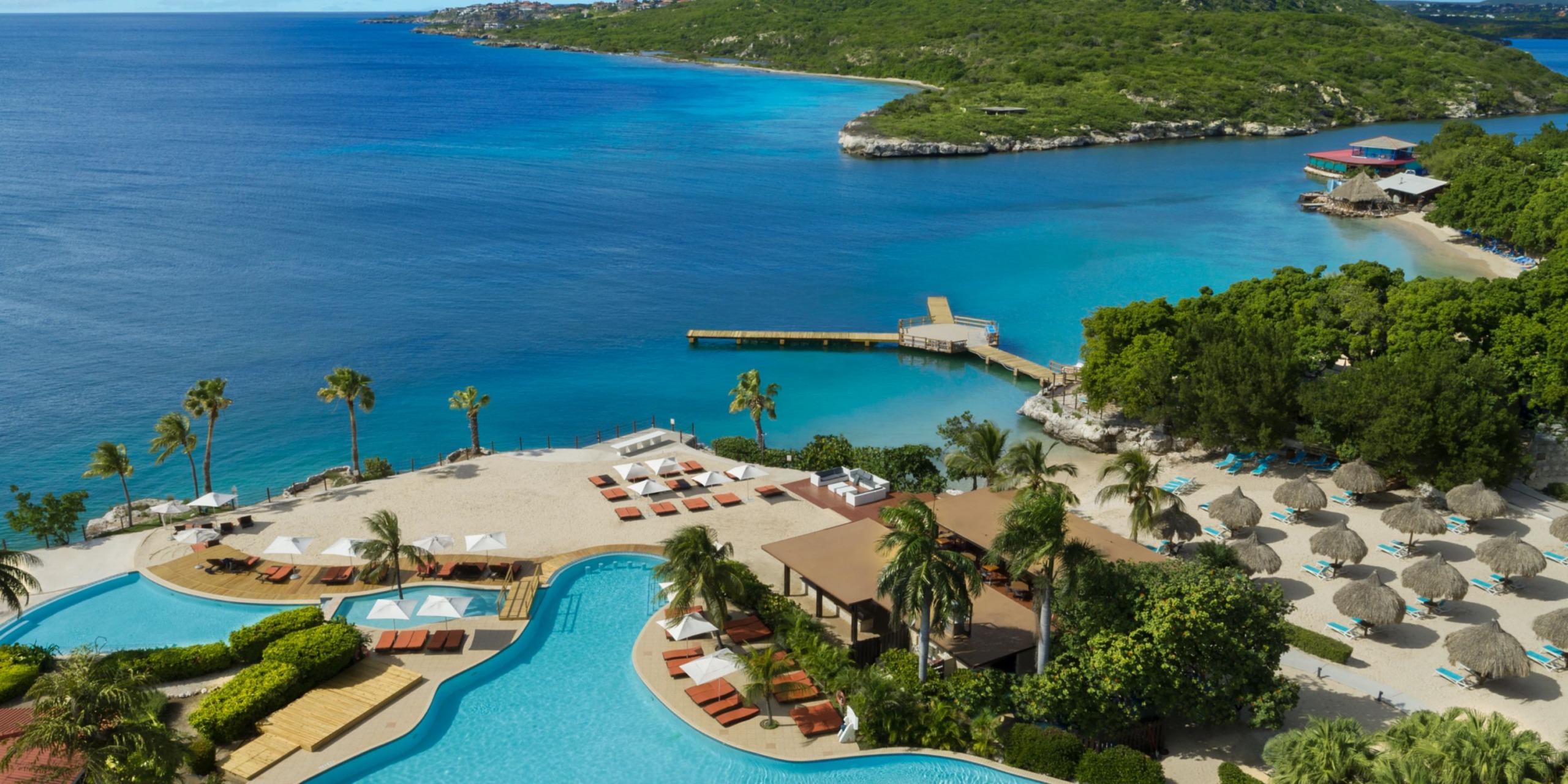 Aerial view of Dreams Curacao, a coastal resort featuring a pool surrounded by lush greenery and sunbeds, with a breathtaking view of the deep blue ocean and hilly island in the background.