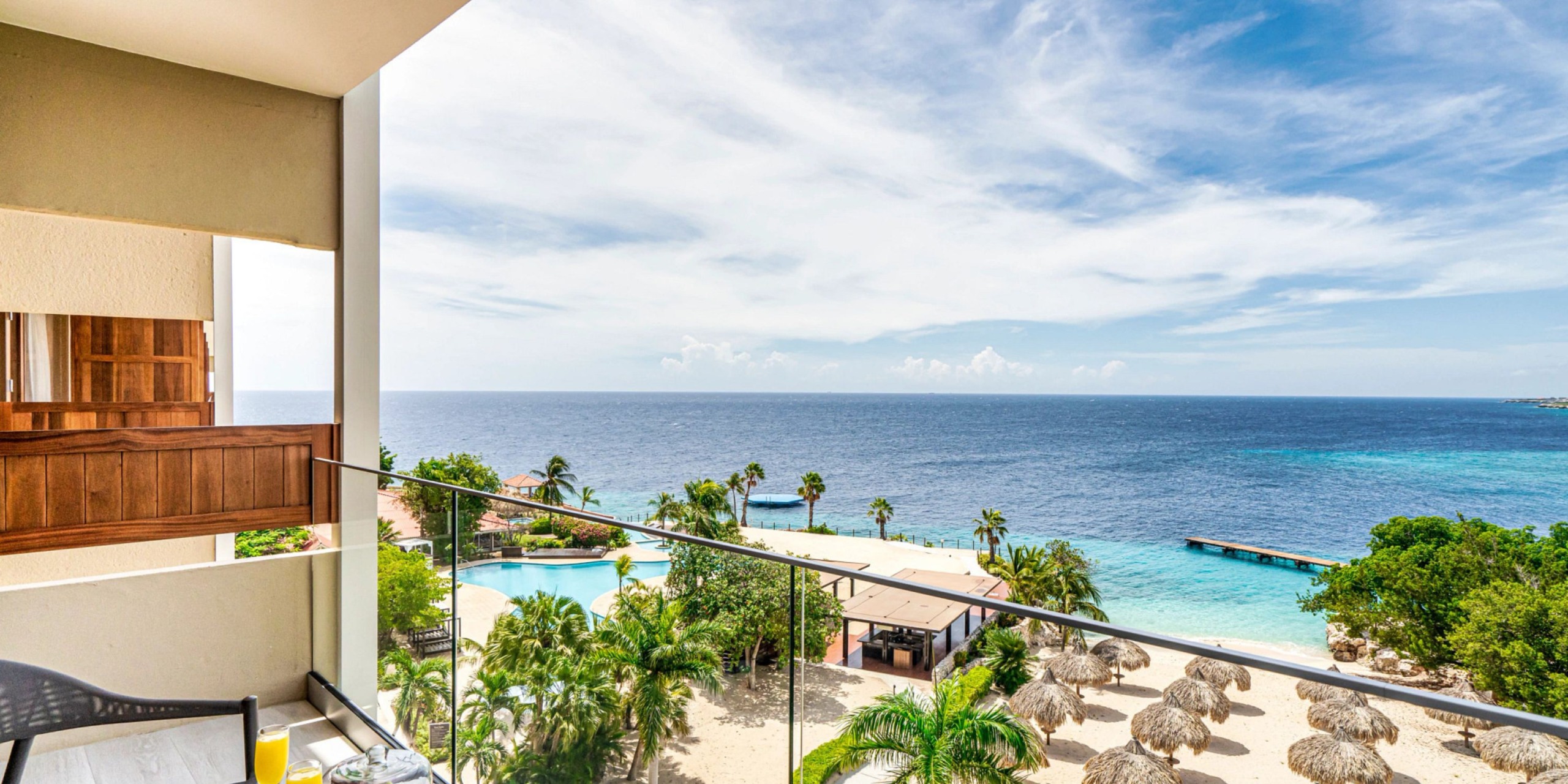 Enjoy the stunning view from a Dreams Curacao balcony, overlooking a tropical beach adorned with palm trees, a refreshing swimming pool, and charming thatched umbrellas. The serene ocean extends to the horizon beneath a partly cloudy sky.