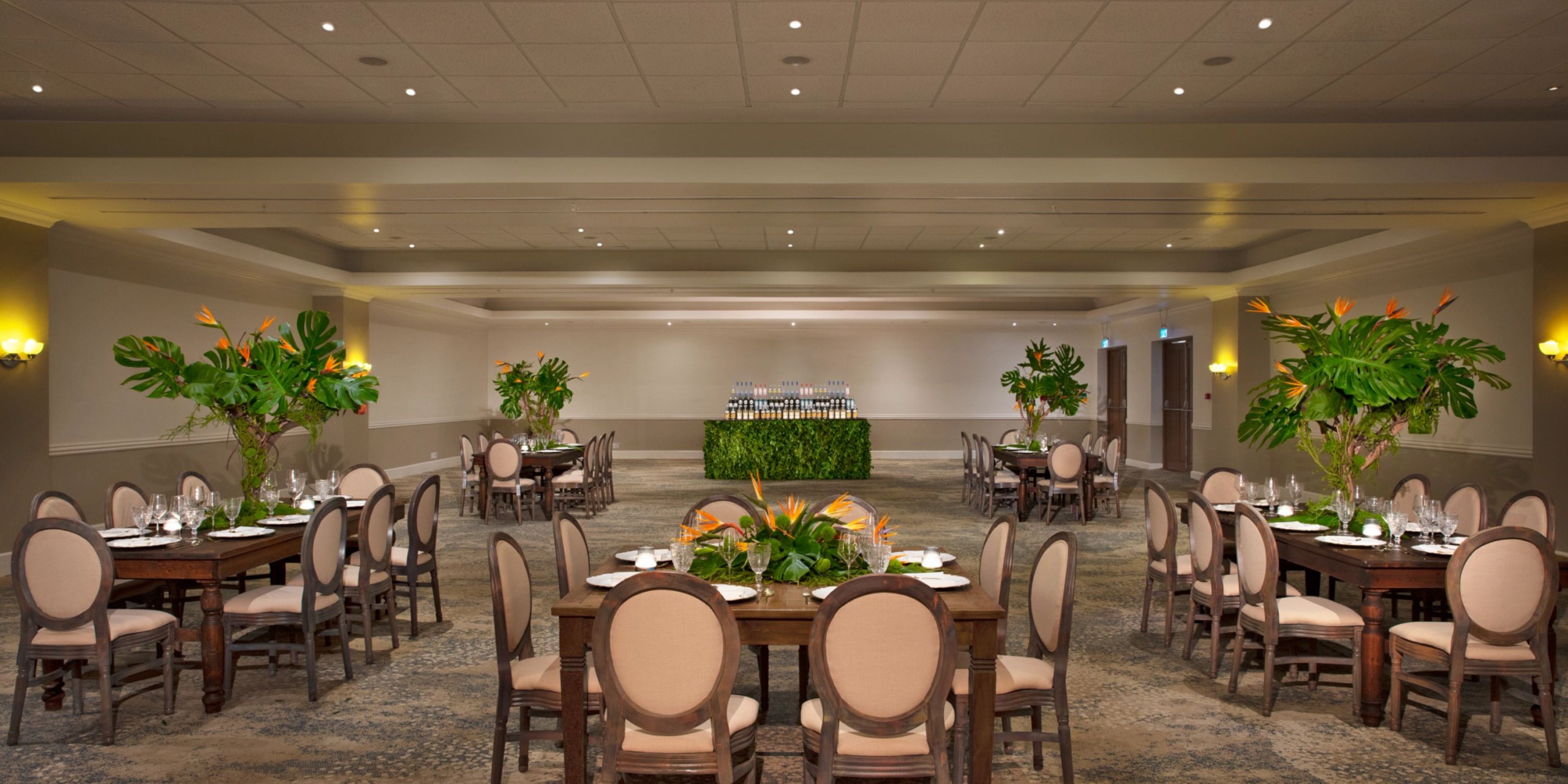The Dreams Curacao banquet hall is spacious and elegantly decorated, featuring round tables with chairs and stunning floral centerpieces. At the back, a table adorned with wine glasses is beautifully nestled among lush plants.