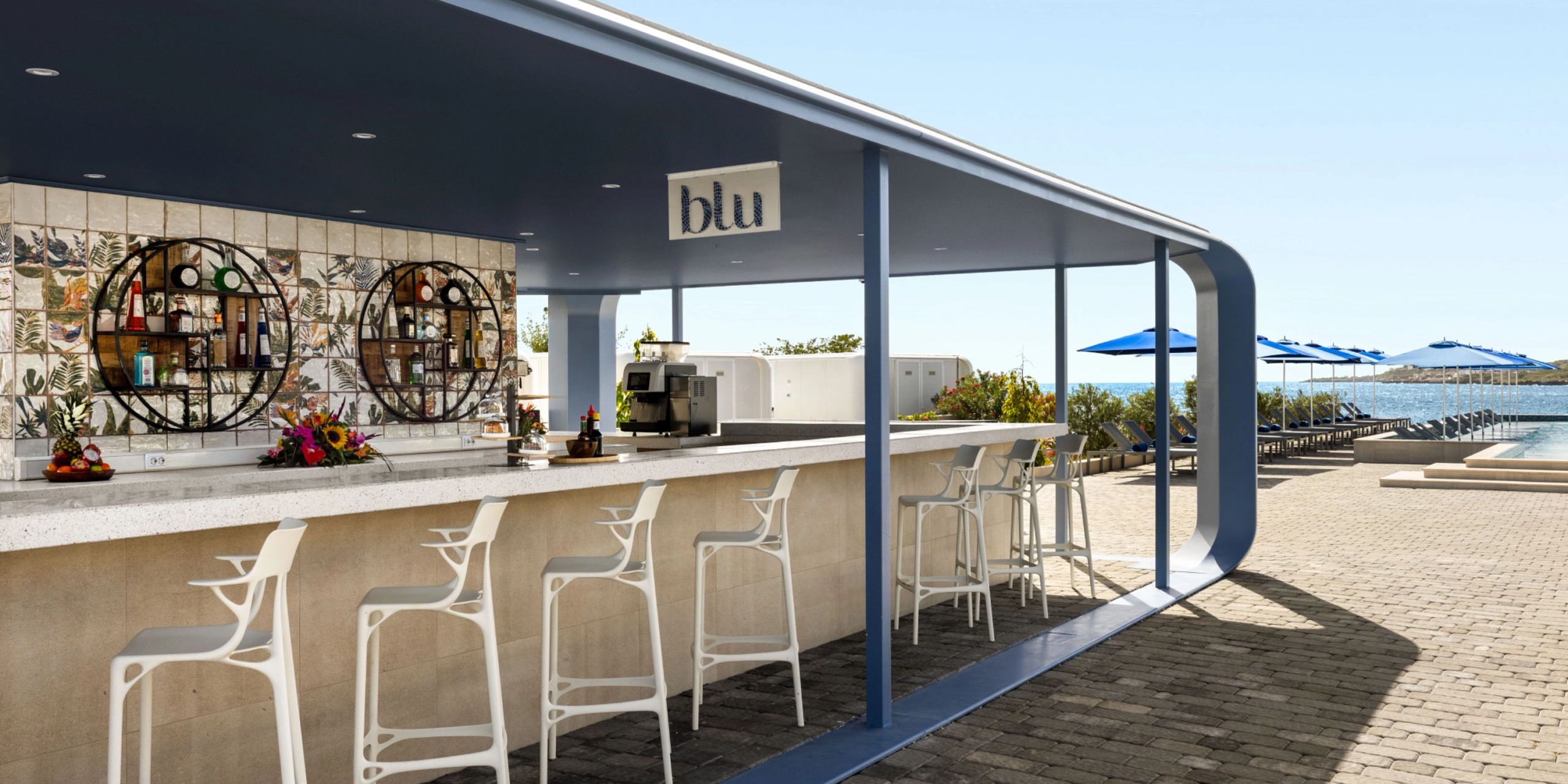 The Dreams Curacao features an outdoor bar complete with stools, a decorative wall behind the counter, and blue umbrellas accompanying a nearby pool in the background.