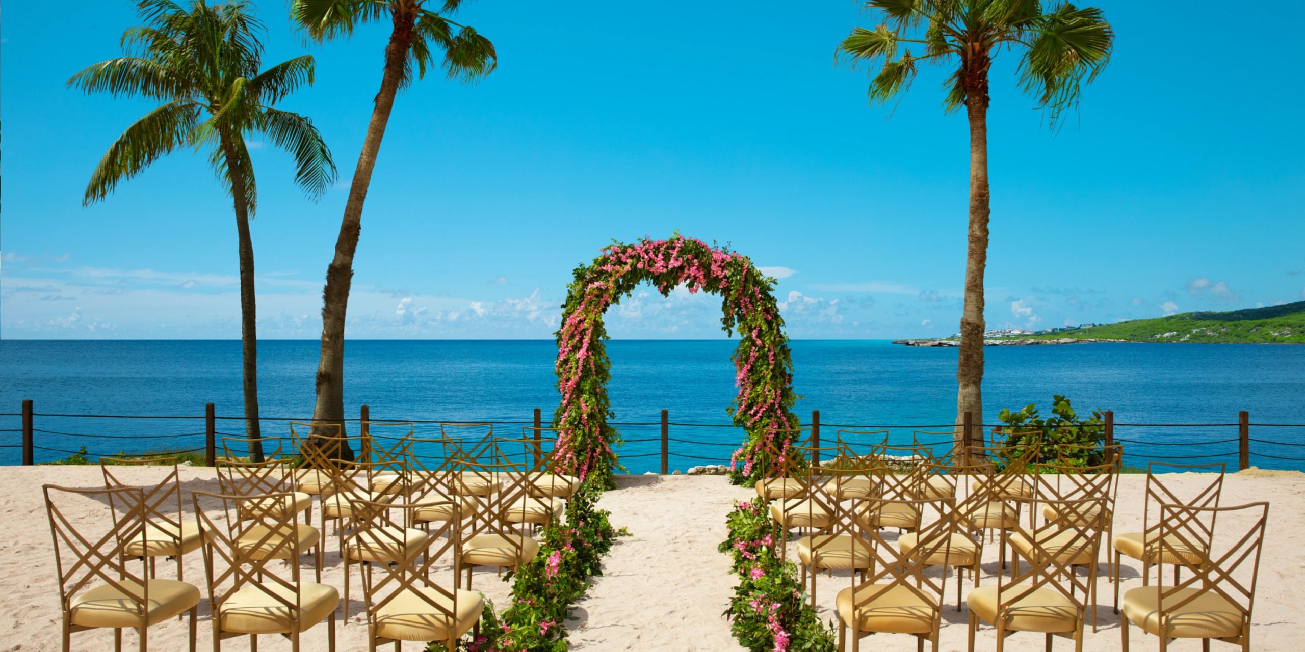 Dreams Curacao offers a beach wedding setup featuring a stunning floral arch and well-organized rows of chairs facing the beautiful ocean, all elegantly surrounded by picturesque palm trees.