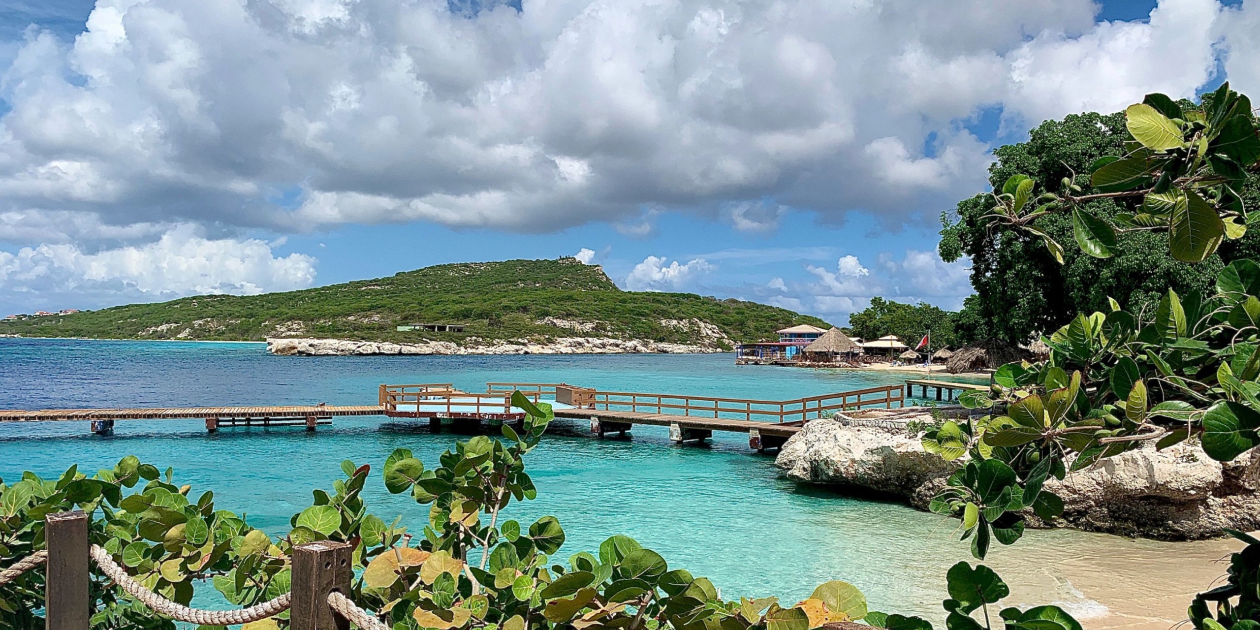 A wooden pier of Dreams Curacao extends over clear turquoise water with a backdrop of lush green hills under a partly cloudy sky.