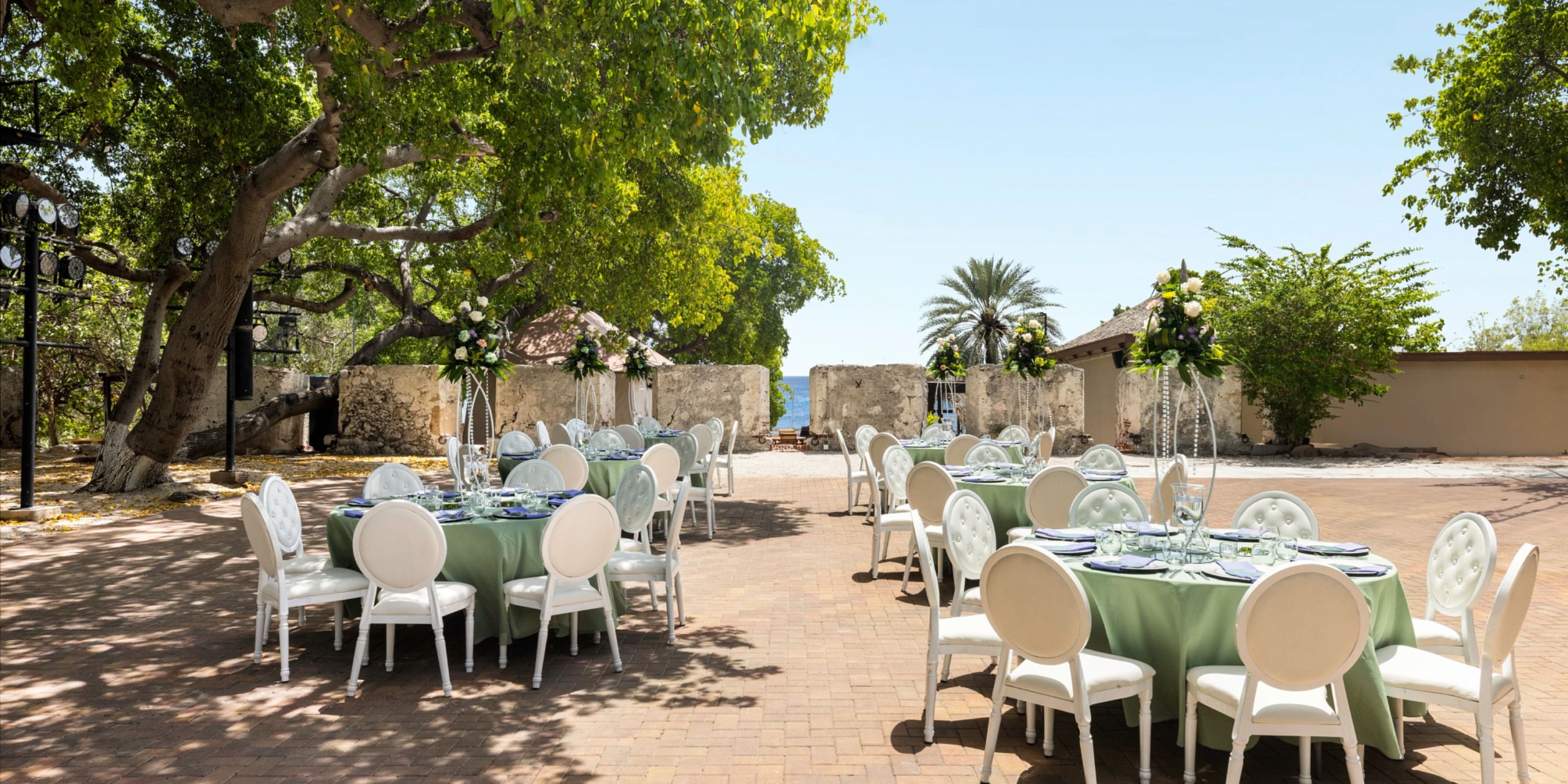 Experience an outdoor event setup at Dreams Curacao, featuring round tables and white chairs on a brick patio, enveloped by trees and open sky.