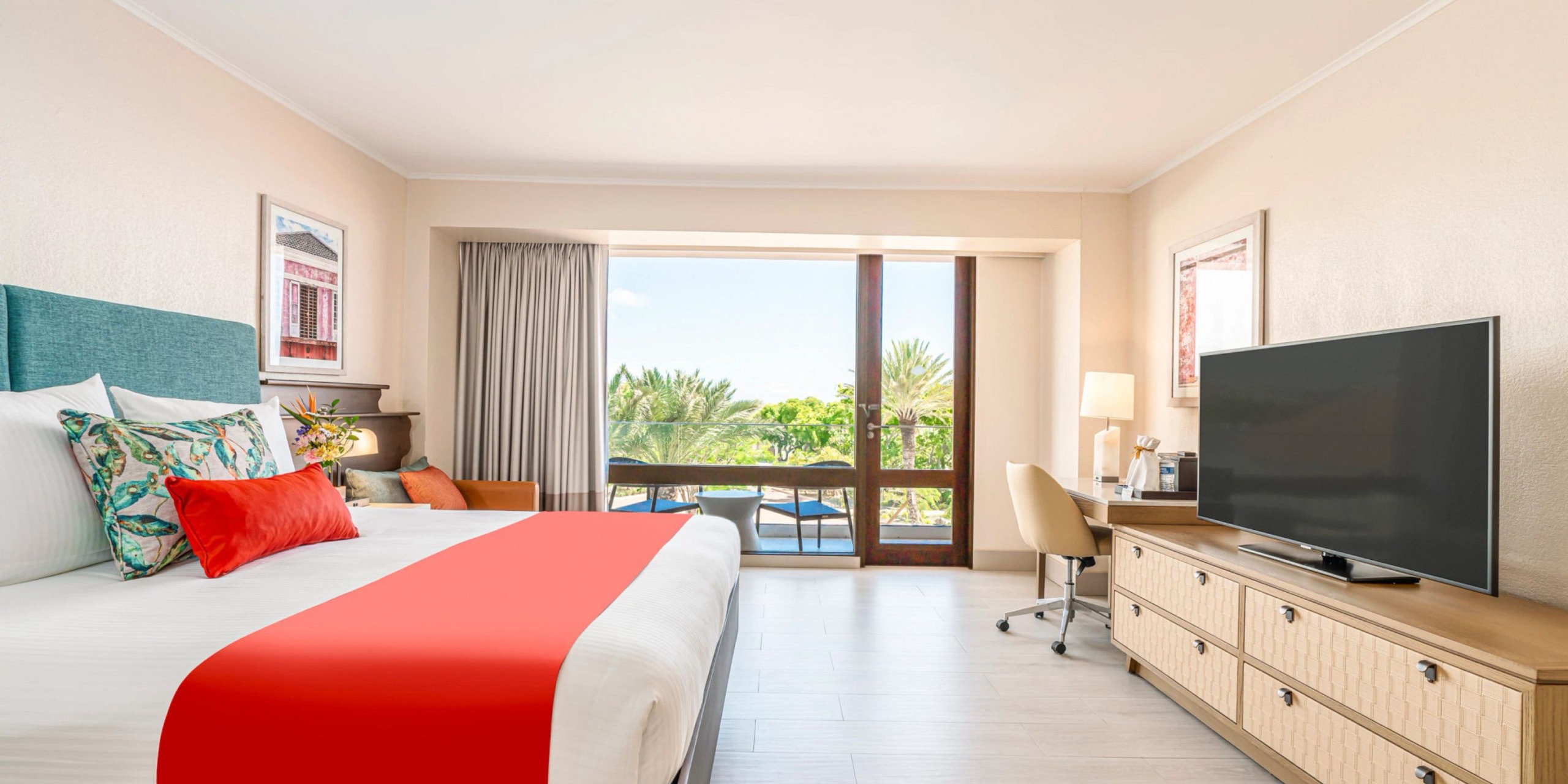 A room at Dreams Curacao features a spacious bed adorned with red and white bedding, a flat-screen TV, a desk paired with a chair, and offers stunning window views of the lush greenery outside.