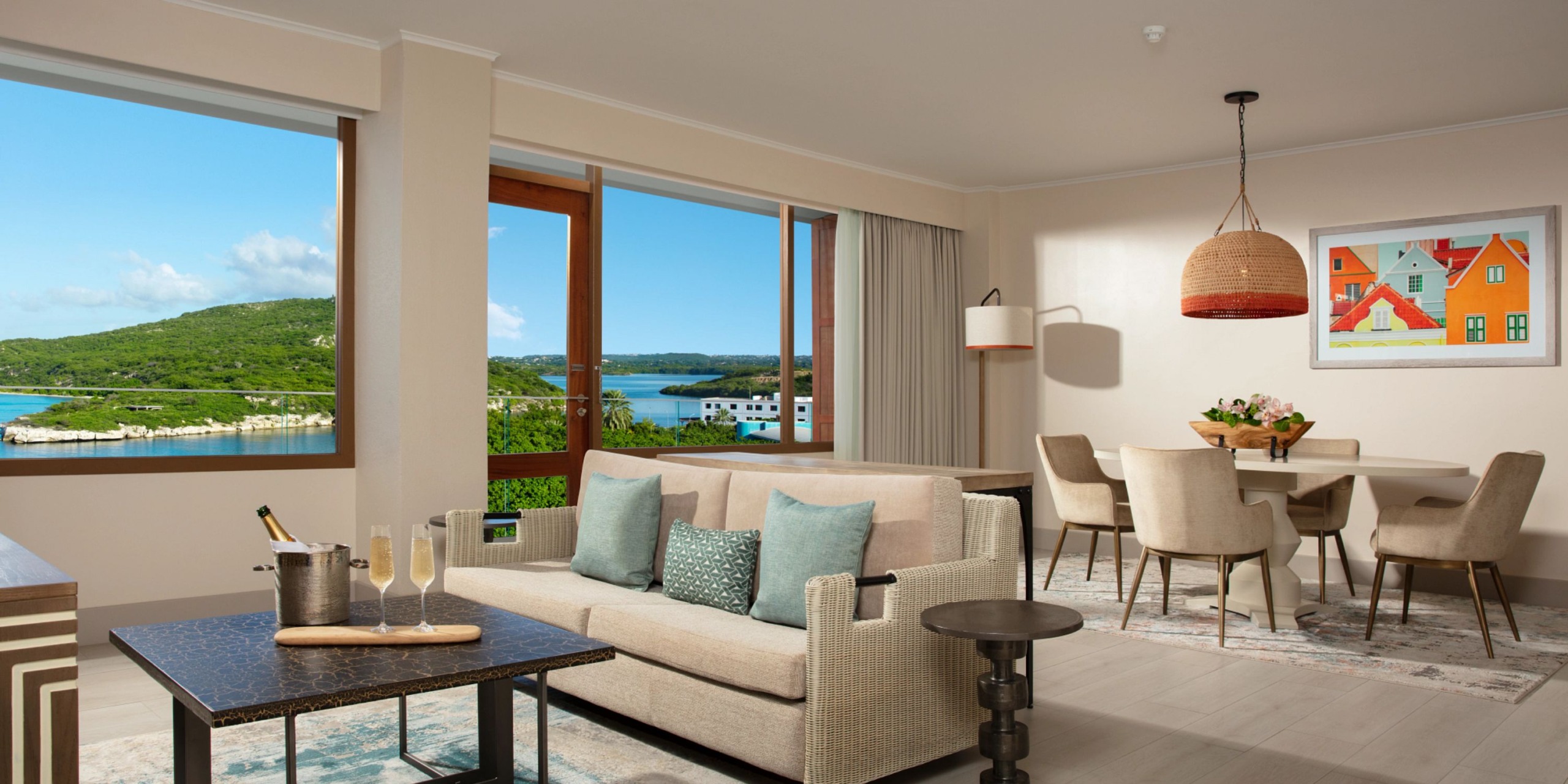 Experience the Dreams Curacao hotel room featuring a seating area, dining table, and expansive windows offering views of a scenic landscape. Adorning the wall are a painting and a pendant light.