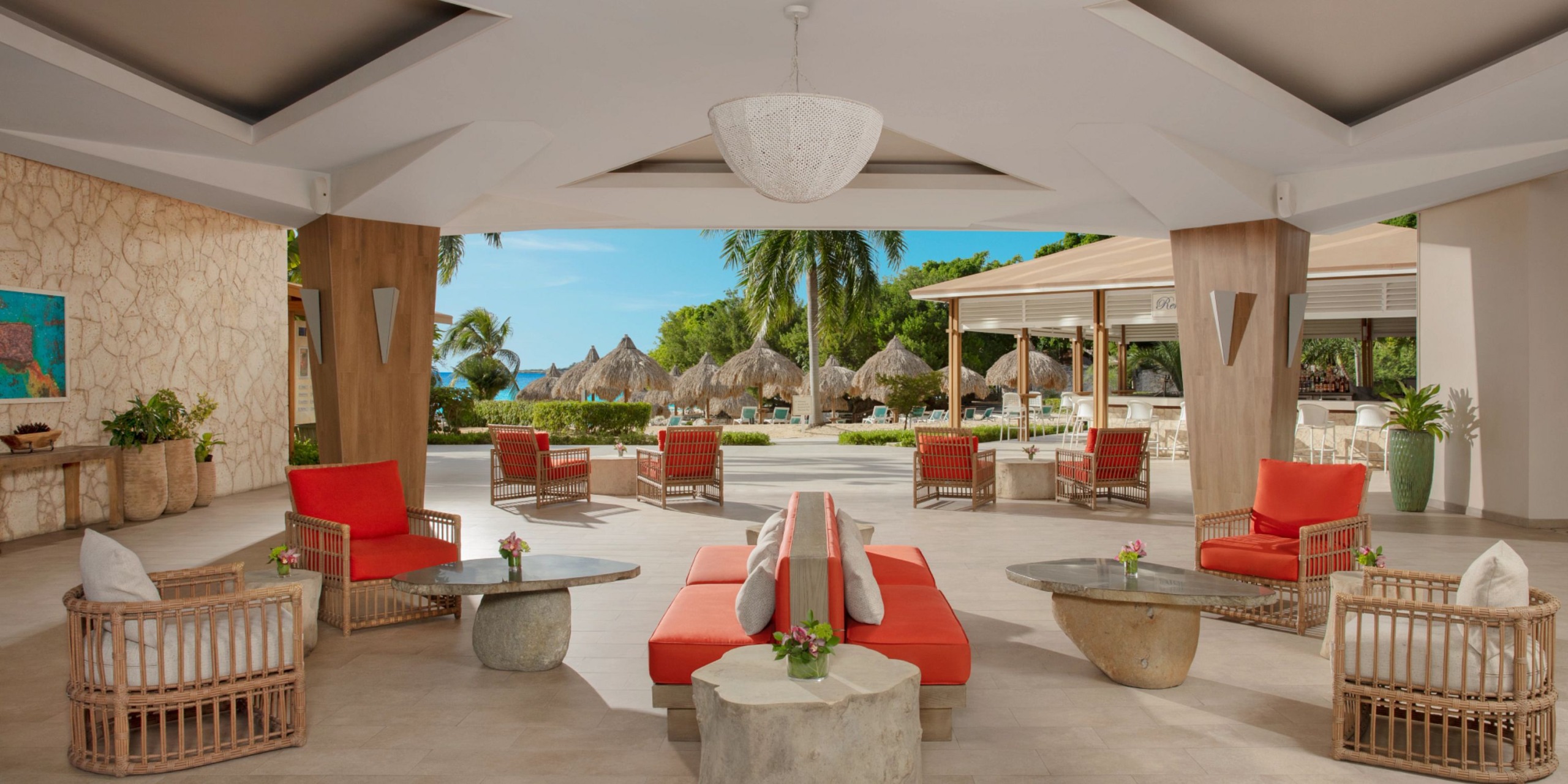 Step into the Dreams Curacao, where you can relax in an open-air lounge area featuring red cushioned seating and stone tables, complemented by a picturesque view of palm trees and thatched umbrellas.