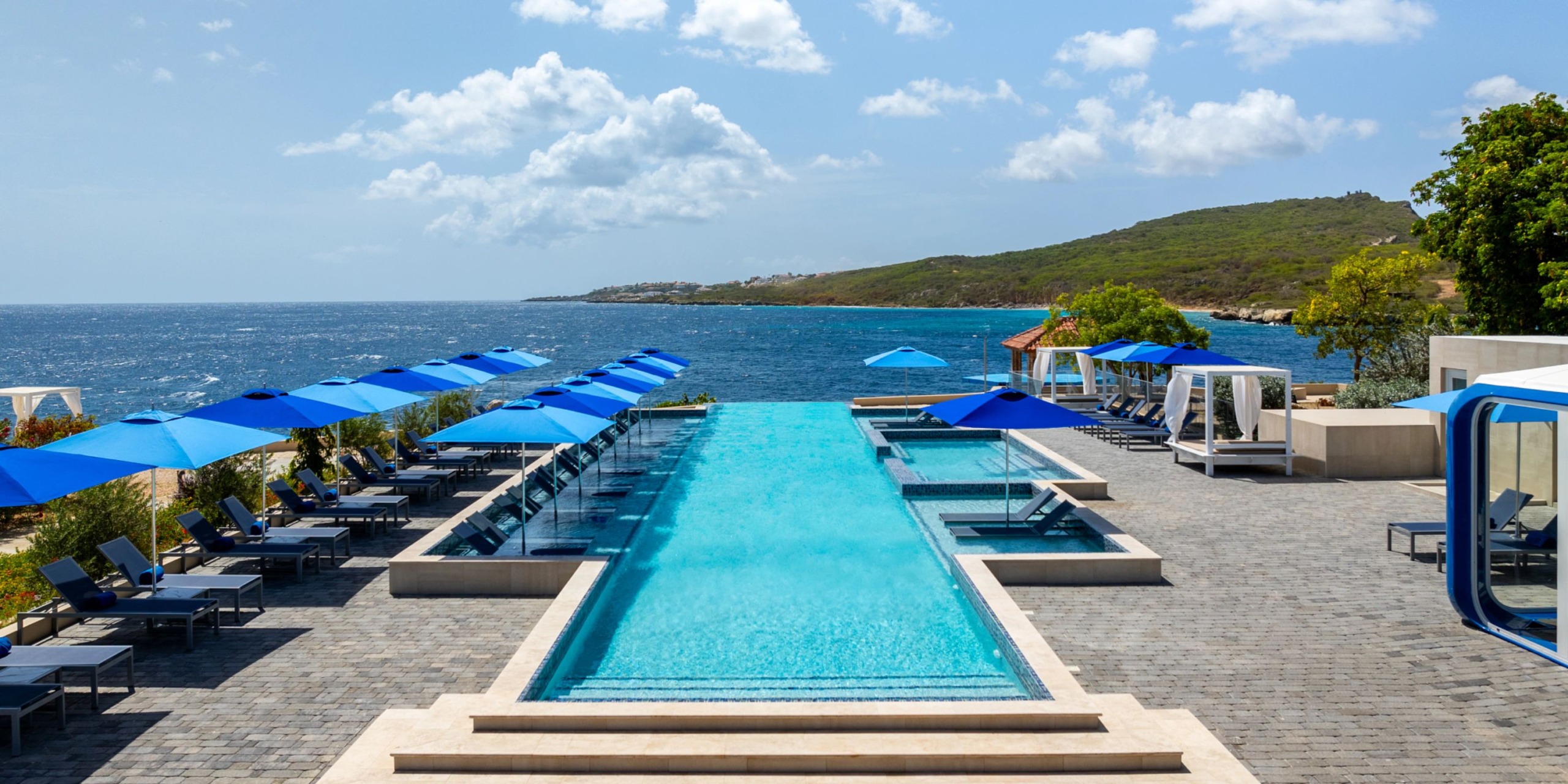 The Dreams Curacao offers an infinity pool with blue umbrellas and lounge chairs that provide stunning views of the ocean, surrounded by lush greenery and rolling hills.