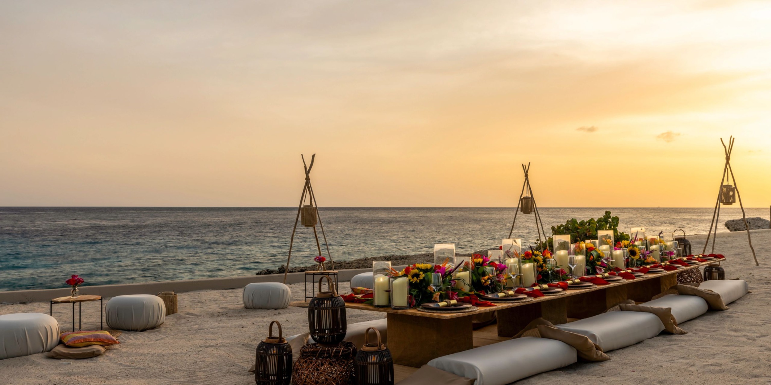 A Dreams Curacao experience features a long table adorned with colorful decorations and pillows, set up on a sandy beach at sunset, overlooking the ocean.