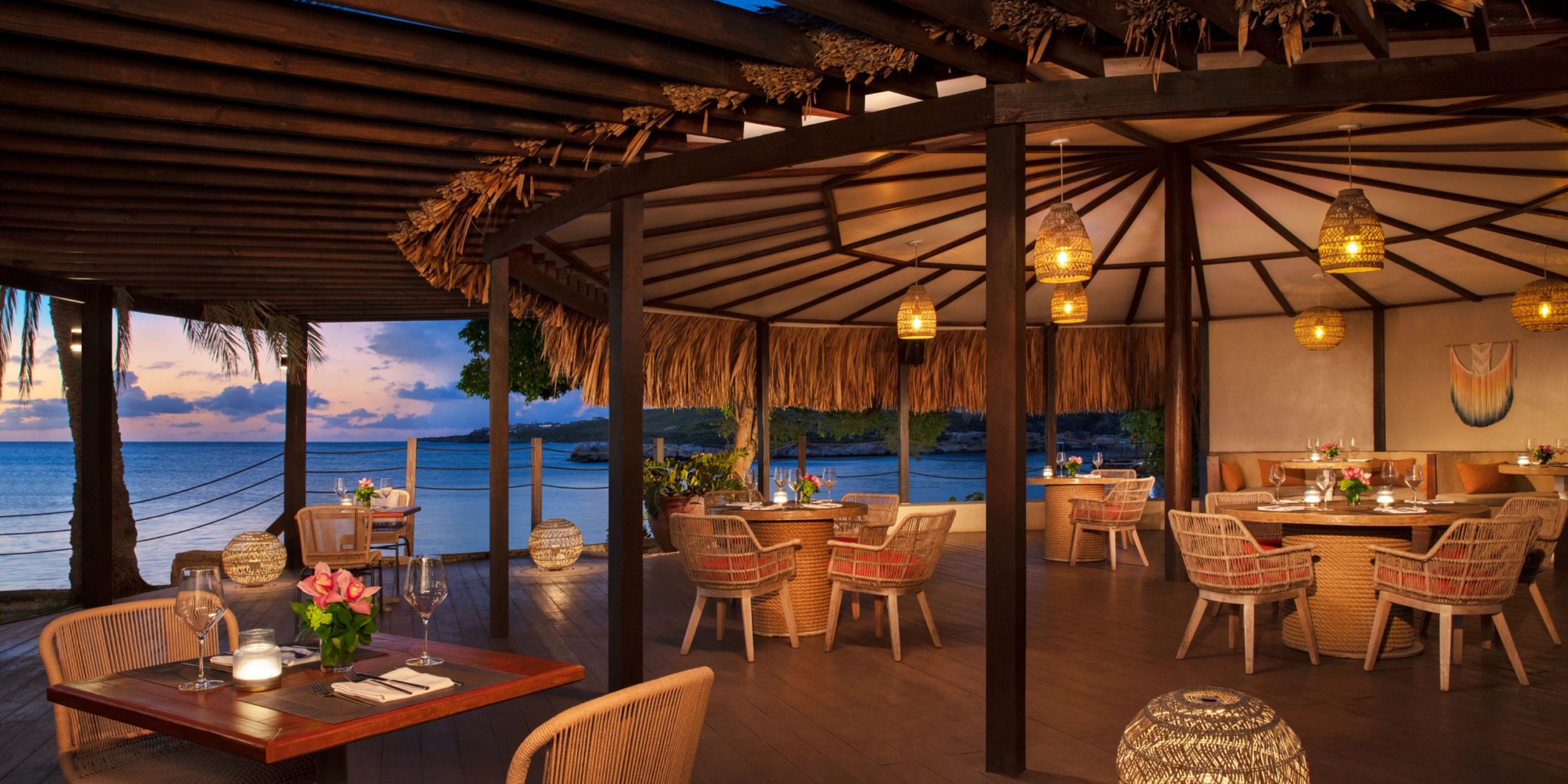 Nestled beneath a thatched roof, Dreams Curacao creates the ultimate outdoor seaside dining experience at sunset, with wooden tables and wicker chairs complemented by decorative lanterns and vibrant floral centerpieces.