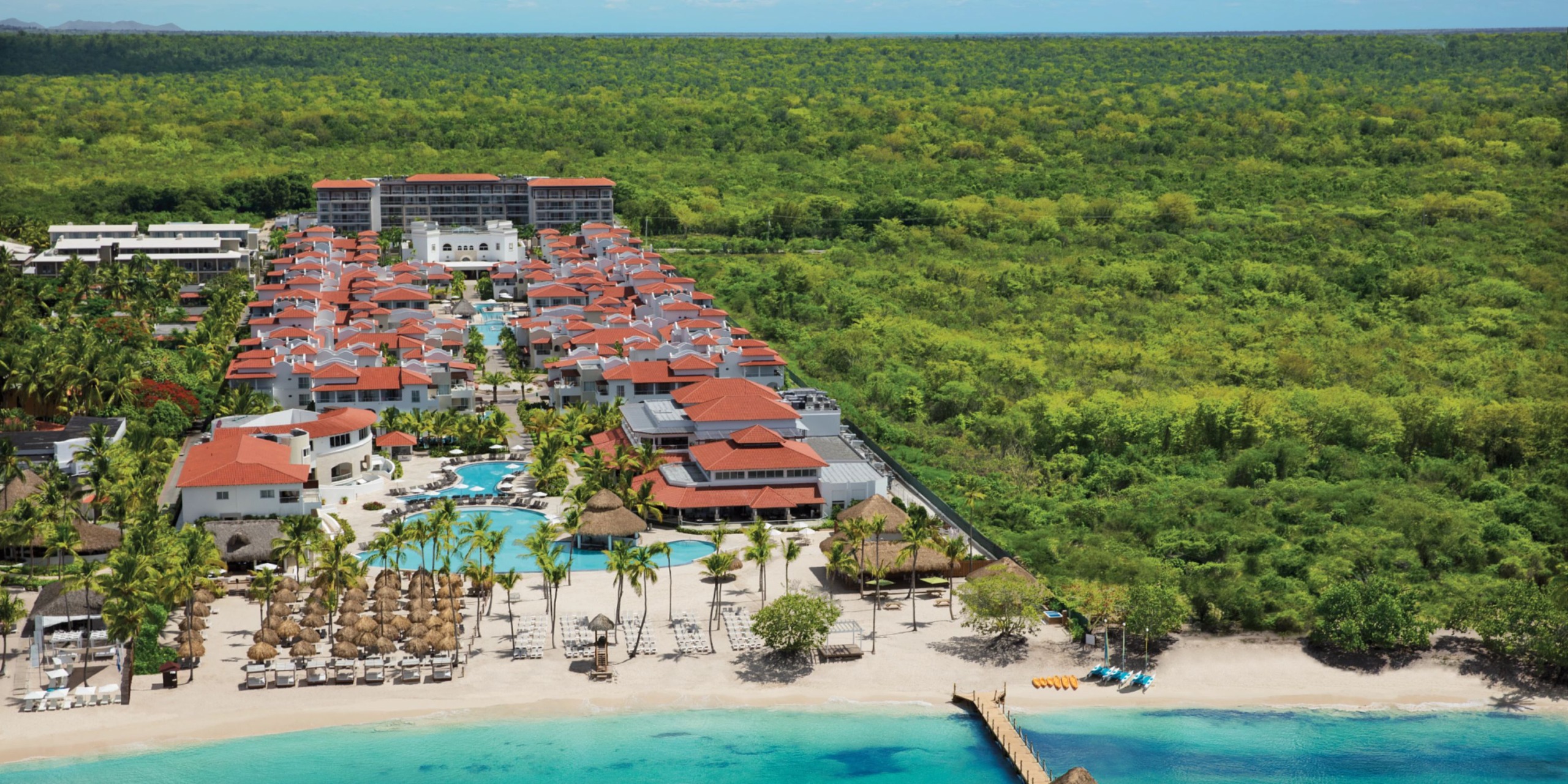 Aerial view of Dreams Dominicus resort showcasing its red-roofed buildings, swimming pools, and palm trees by a beach with turquoise water, surrounded by lush green forest.