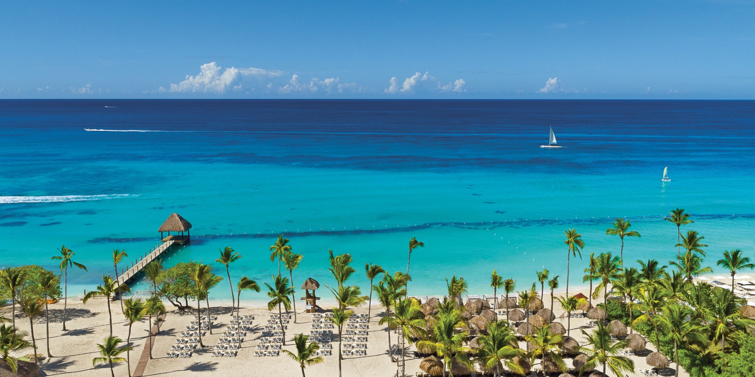 The scene features Dreams Dominicus, with its tropical beach adorned by palm trees, clear turquoise waters, a wooden pier stretching out to sea, and a sailboat gracefully sailing on the horizon beneath a vibrant blue sky.