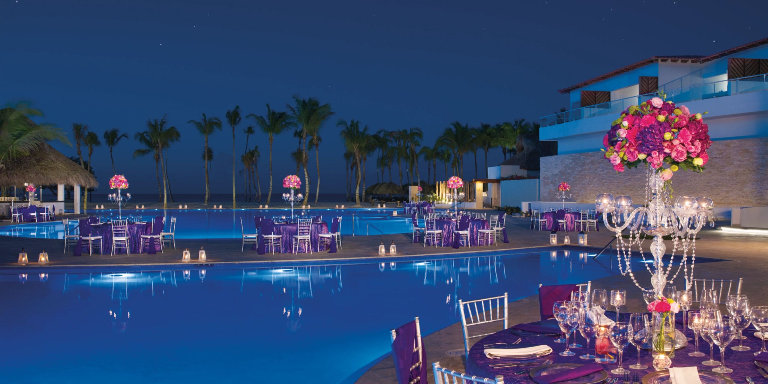 Experience a sophisticated outdoor dining scene at Dreams Dominicus, set by a large pool. Enjoy tables adorned with purple tablecloths, decorative crystal candelabras, and vibrant pink floral arrangements, all under the serene night sky and surrounded by majestic palm trees.
