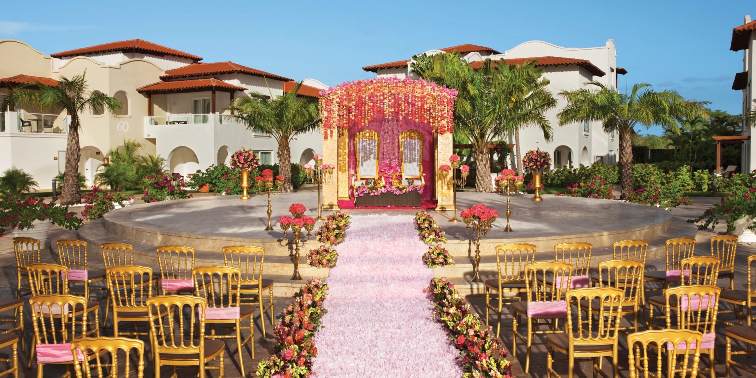 Outdoor wedding setup featuring the Dreams Dominicus with a pink flower-decorated mandap, surrounded by golden chairs and palm trees, set against a backdrop of Mediterranean-style buildings.