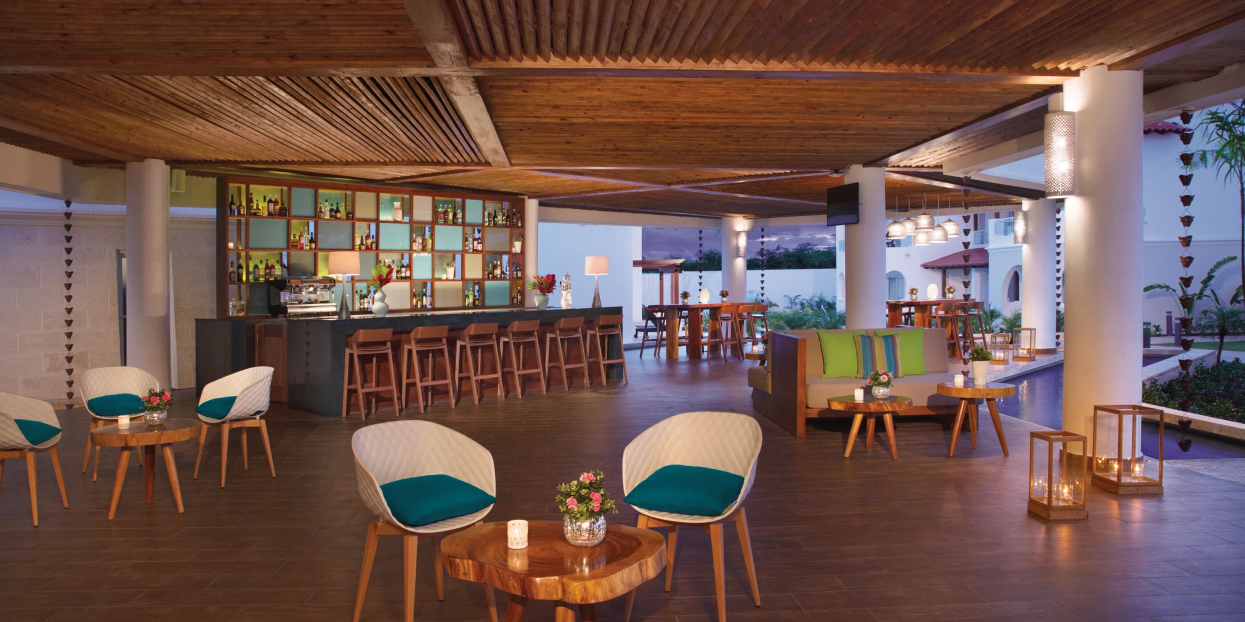 Experience the Dreams Dominicus outdoor lounge, featuring a wooden ceiling, a bar equipped with stools, round tables accompanied by chairs, and beautifully arranged decorative plants.