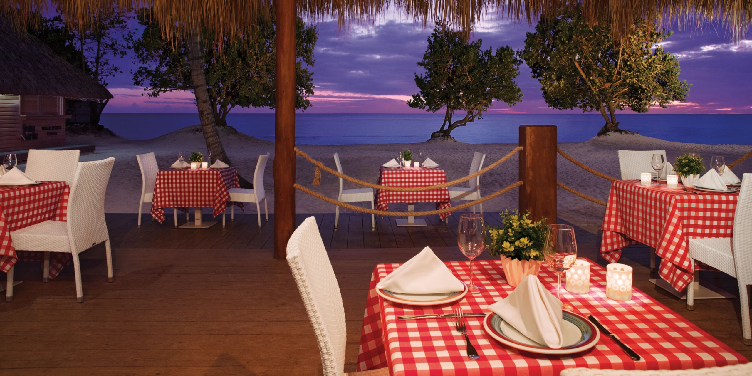 Dreams Dominicus offers an outdoor beachside dining experience at sunset, complete with tables adorned with red checkered tablecloths, white chairs, and a stunning ocean view.