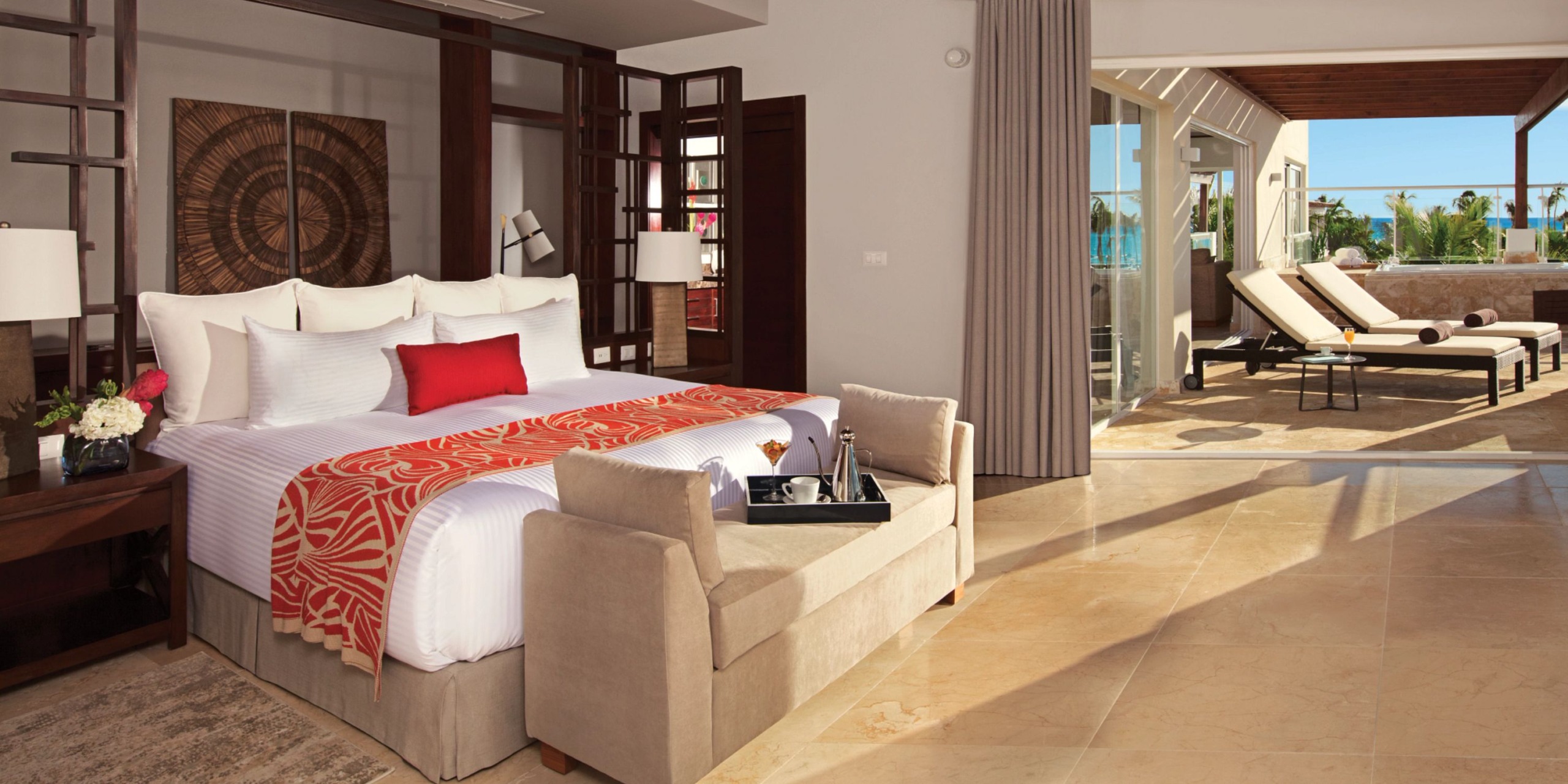 A luxurious bedroom at Dreams Dominicus showcases a king-size bed adorned with red and white decor, a cozy sitting area, and large windows that open to a sunny terrace equipped with lounge chairs.