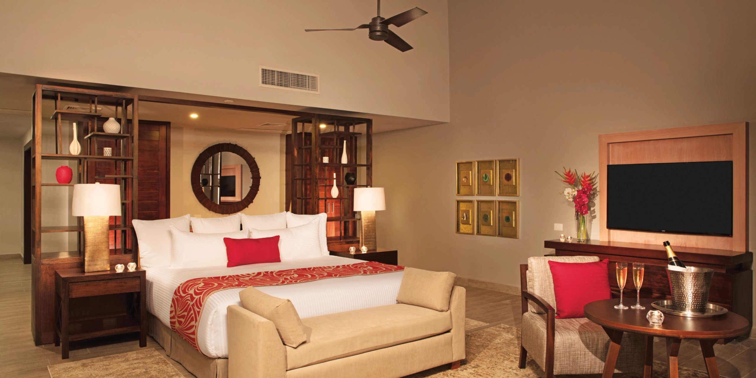 The Dreams Dominicus features a contemporary room with a spacious bed highlighted by red accents, a cozy seating area, a wall-mounted television, chic decor, and a ceiling fan.