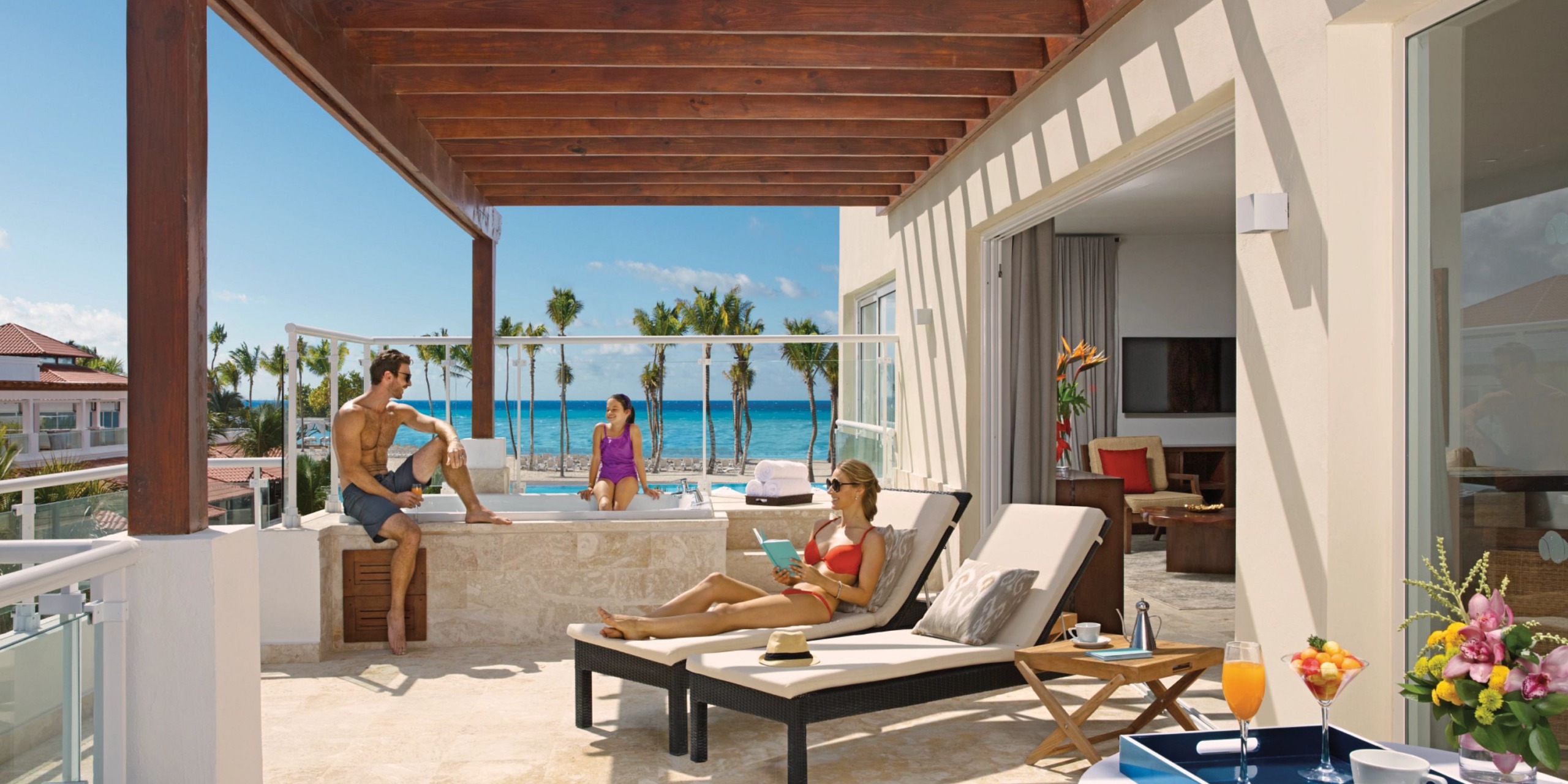 A group of people unwind on a terrace featuring Dreams Dominicus lounge chairs and a hot tub, all with a view of the beach.