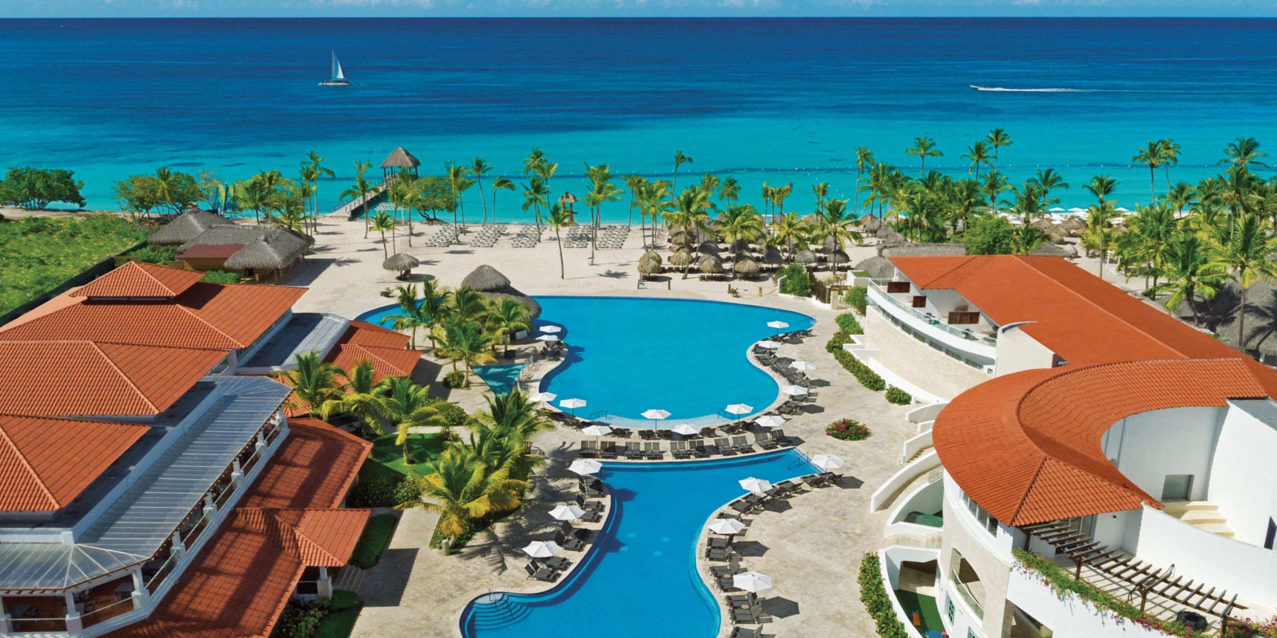Aerial view of Dreams Dominicus, a beachfront resort featuring a large pool, sun loungers, and palm trees, overlooking the blue ocean and sandy beach.
