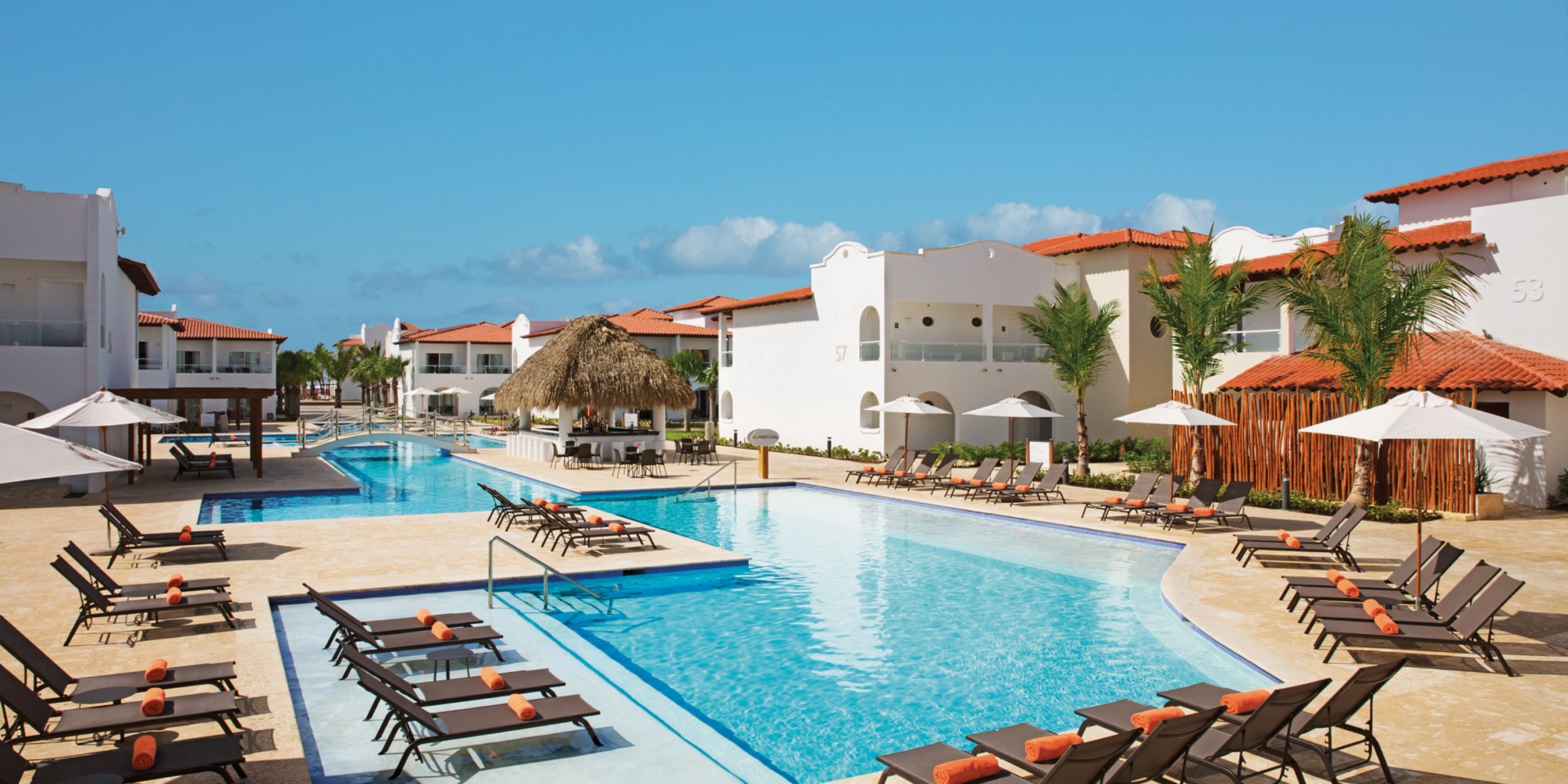 Discover the Dreams Dominicus resort's luxurious pool area, complete with comfortable lounge chairs and umbrellas, surrounded by white buildings featuring charming red-tiled roofs beneath a crystal-clear blue sky.