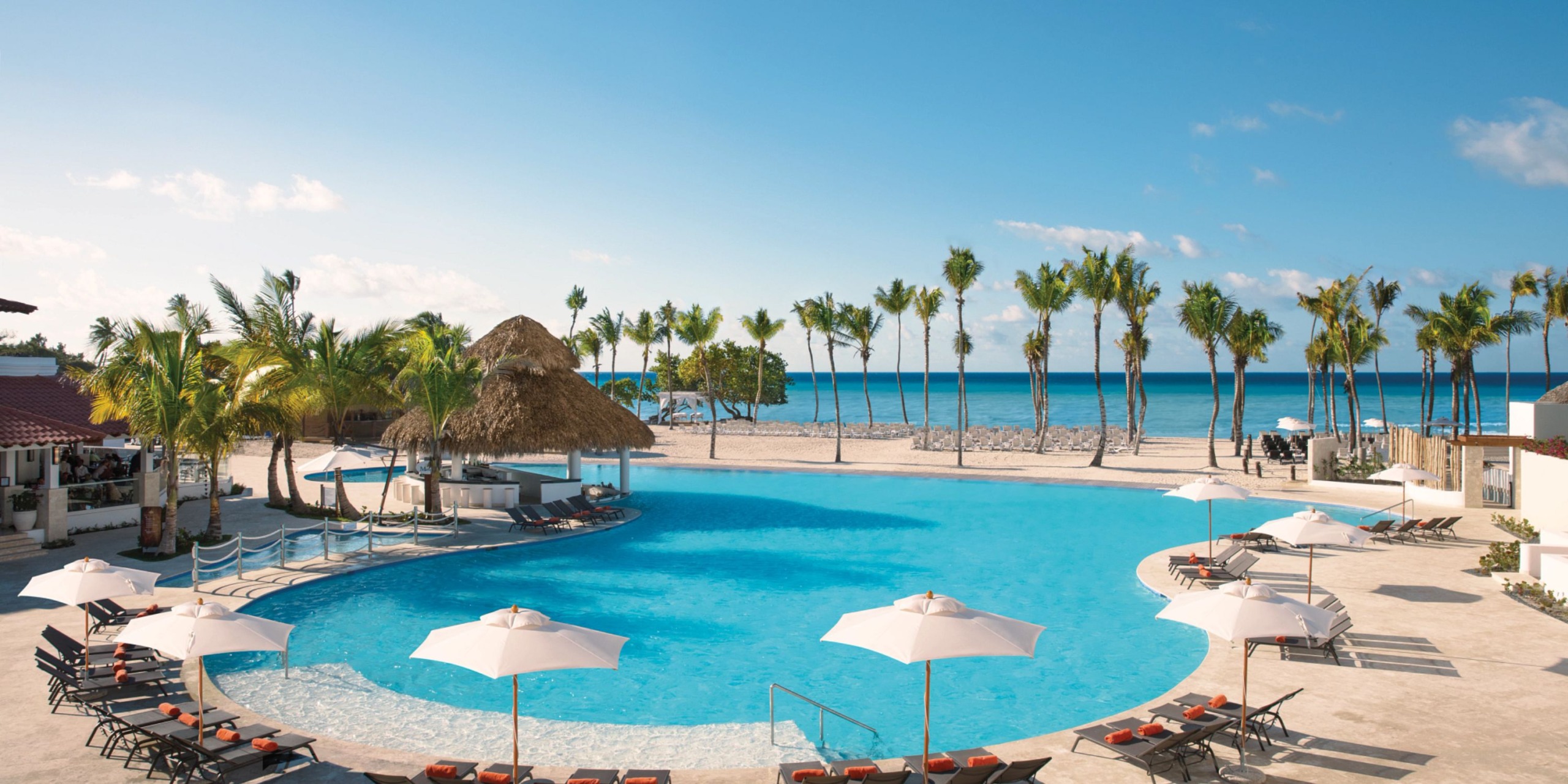 At Dreams Dominicus, the pool area, adorned with palm trees, loungers, and umbrellas, offers a splendid view of a sandy beach and the ocean beneath a clear blue sky.