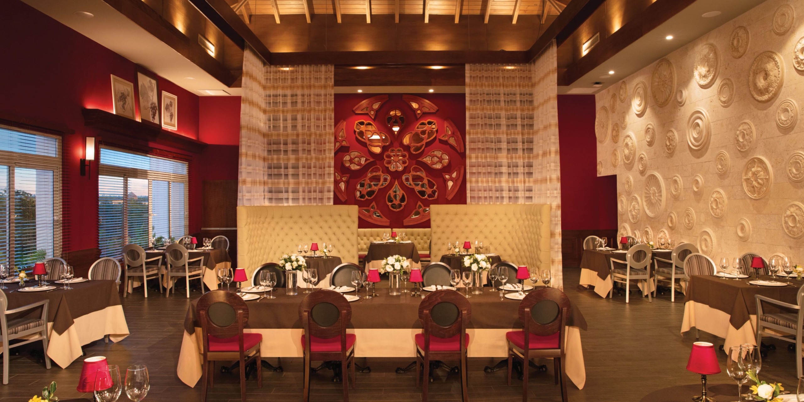 The Dreams Dominicus boasts an elegant interior with red walls, intricately patterned panels, and ornate decor. The tables are adorned with white tablecloths and flowers, accompanied by chairs featuring red cushions. Warm lighting envelops the space, creating a cozy atmosphere.