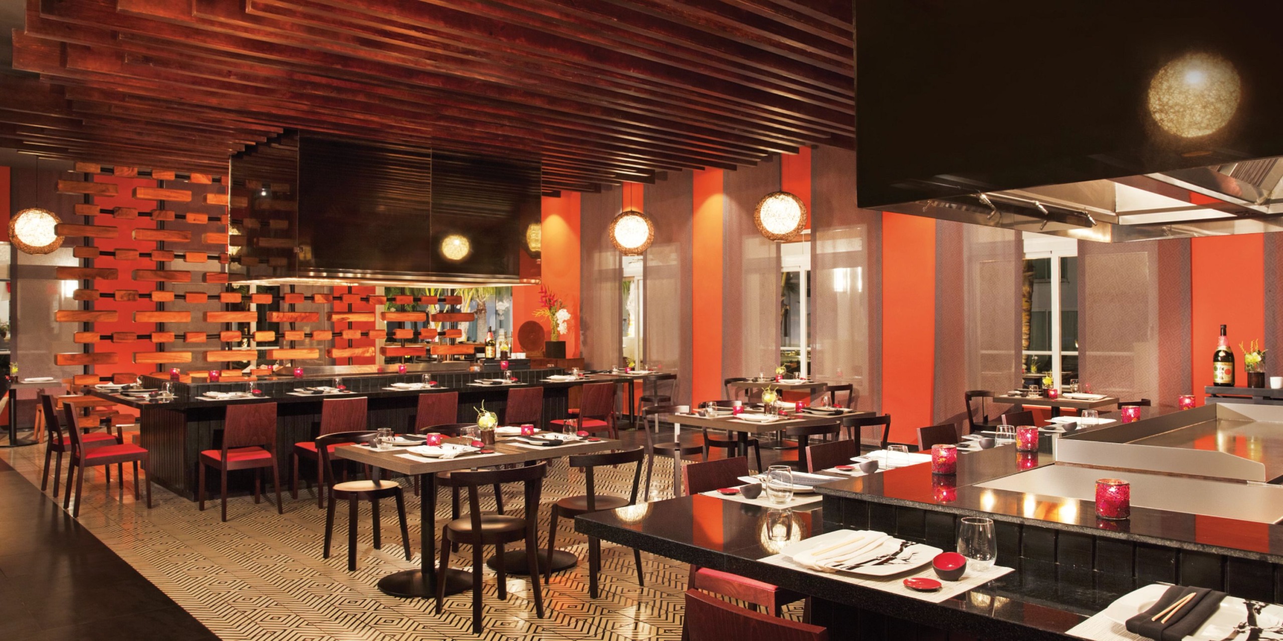 The Dreams Dominicus boasts a modern restaurant interior featuring wooden accents, red and black chairs, tables elegantly set for dining, and an open kitchen area. Soft lighting enhances the cozy atmosphere.