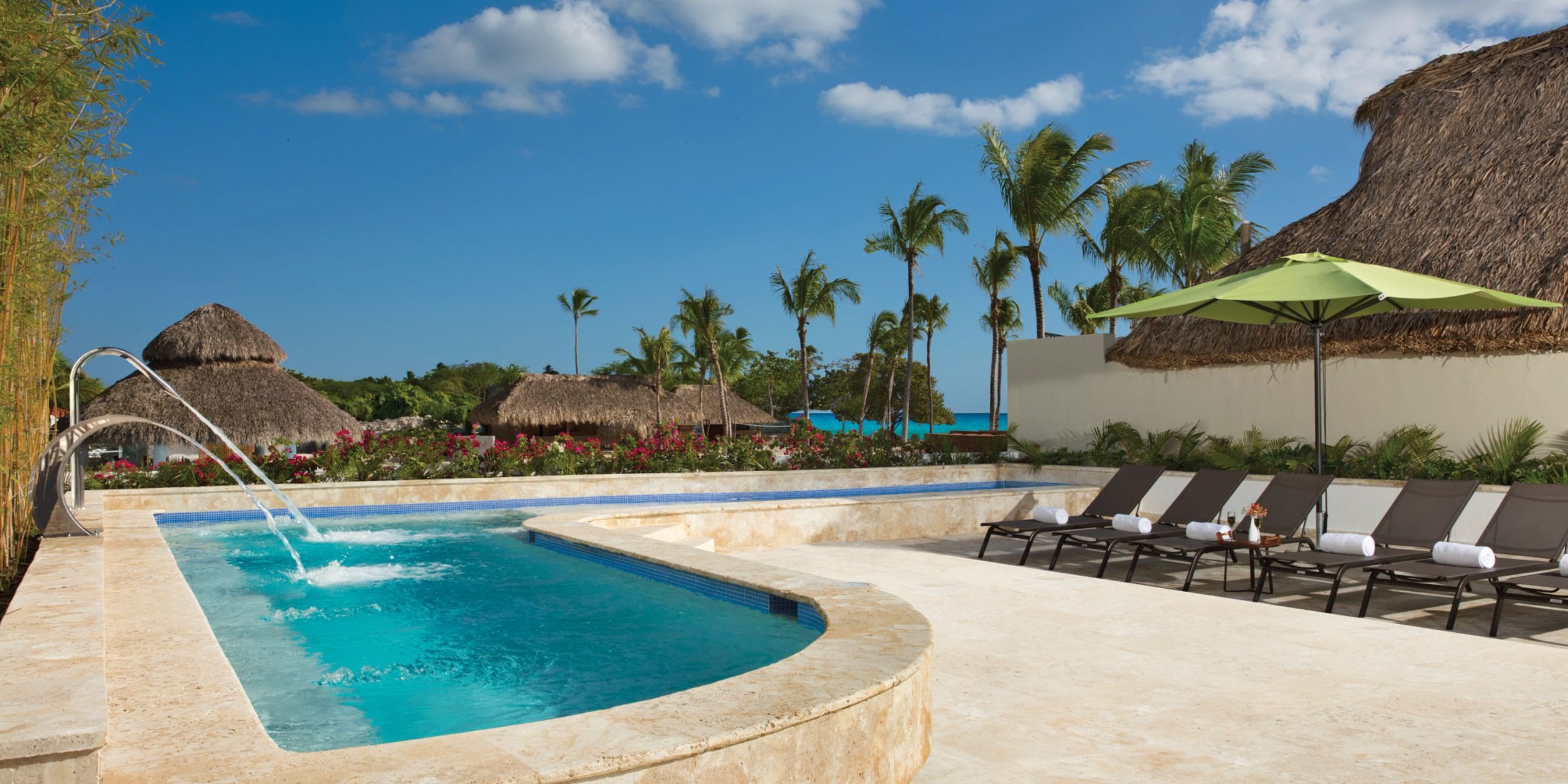 The Dreams Dominicus resort features a luxurious pool adorned with cascading water features, encircled by lounge chairs and vibrant green umbrellas. In the background, majestic palm trees and charming thatched buildings stand under a pristine blue sky.