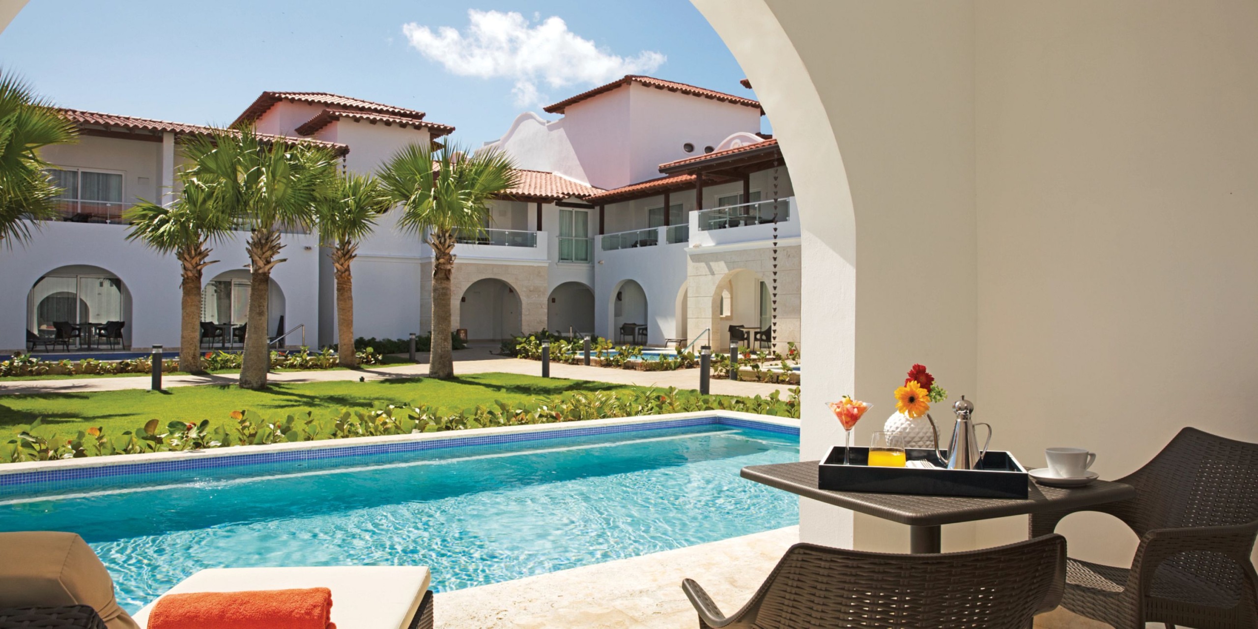 A patio view of Dreams Dominicus resort showcases a private pool, palm trees, and multi-story buildings with arched balconies. In the foreground, there is a table adorned with drinks and flowers.