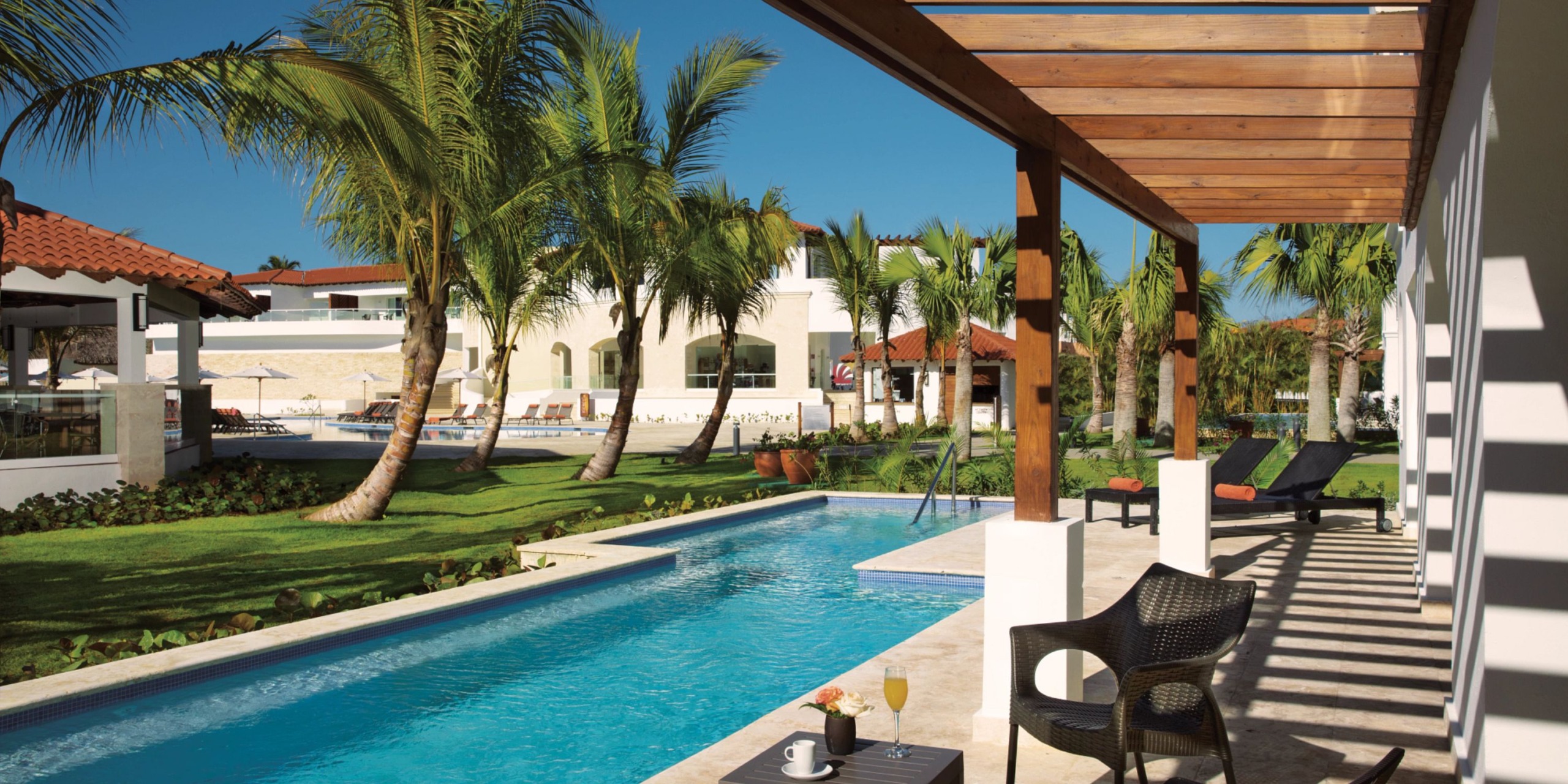 A tranquil swimming pool next to a Dreams Dominicus patio set featuring chairs and a small table. Palm trees and lush greenery envelop the area, with buildings visible in the background against a clear blue sky.