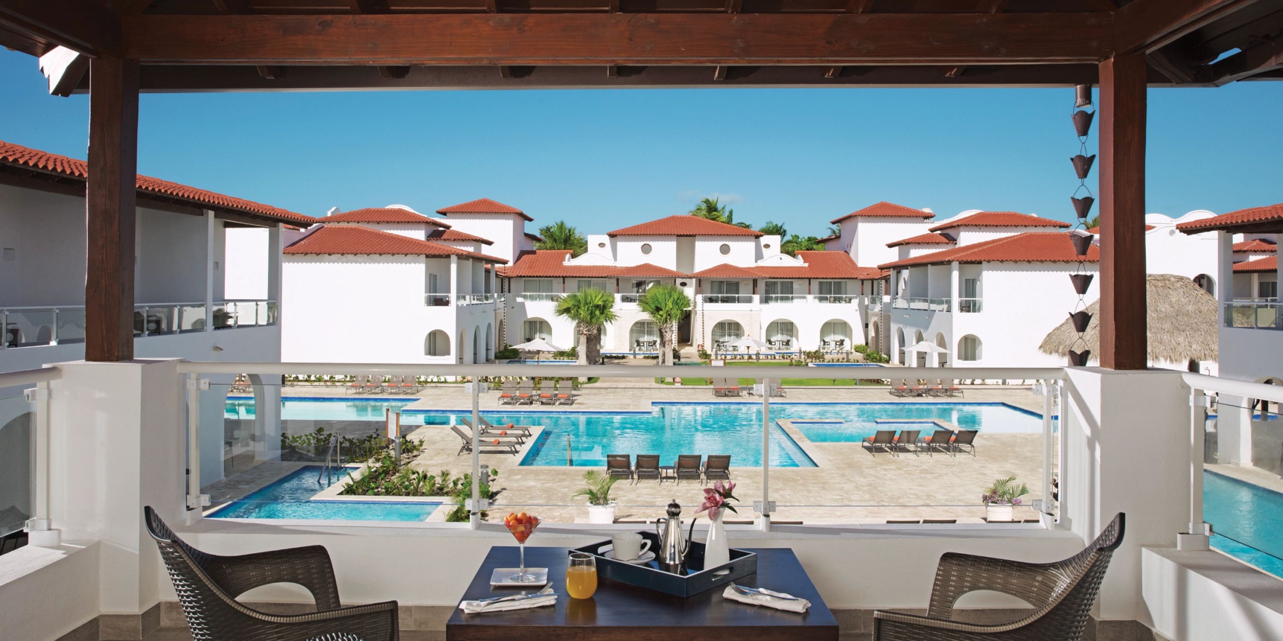 A view of Dreams Dominicus featuring a pool, charming white buildings with red roofs, and comfortable lounge chairs. In the foreground, a table adorned with drinks and flowers is situated under a wooden pergola.