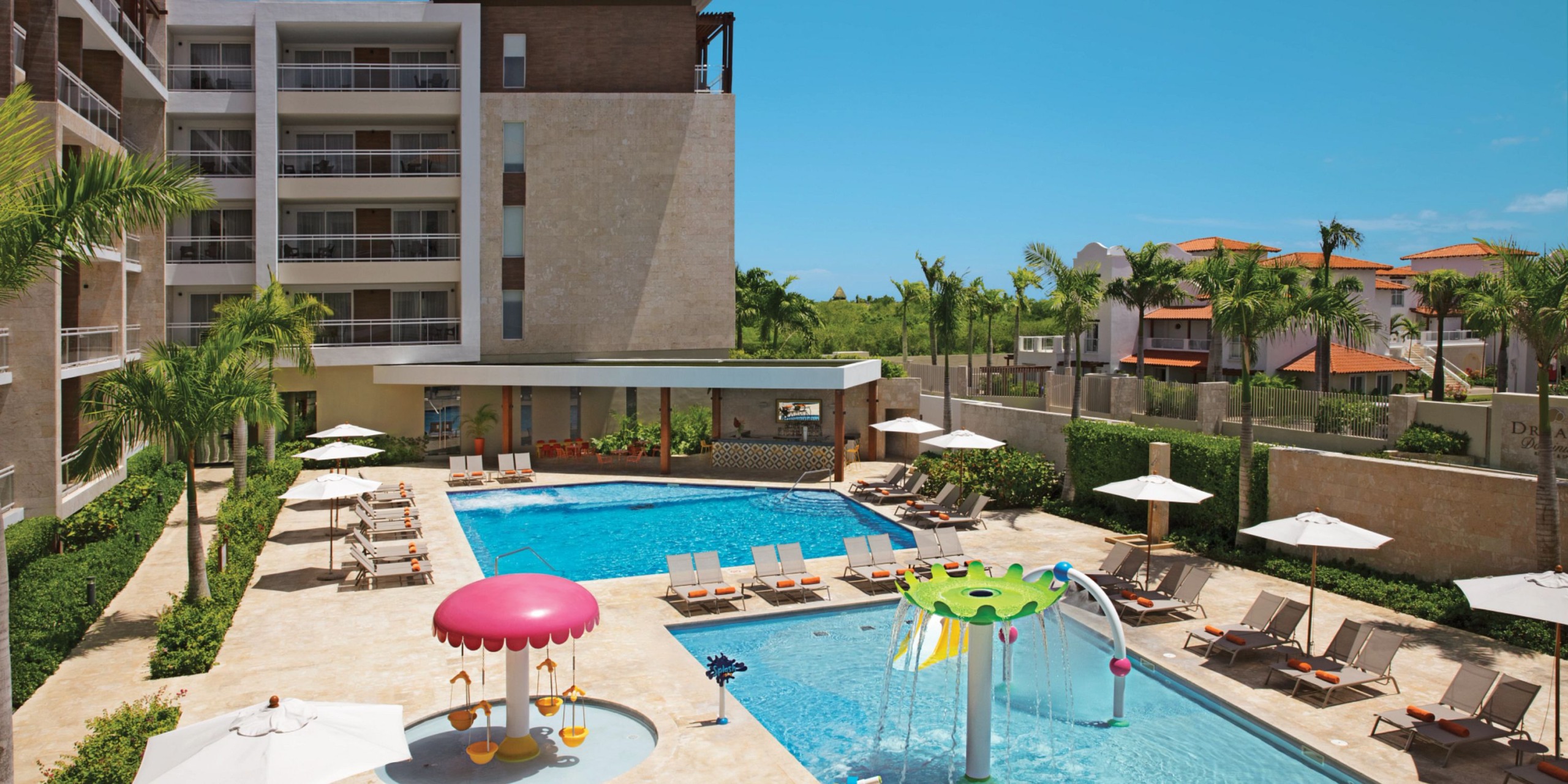 View of the Dreams Dominicus swimming pool area, complete with lounge chairs, umbrellas, and a kids' splash zone showcasing colorful water features, all nestled among lush greenery and surrounding buildings.