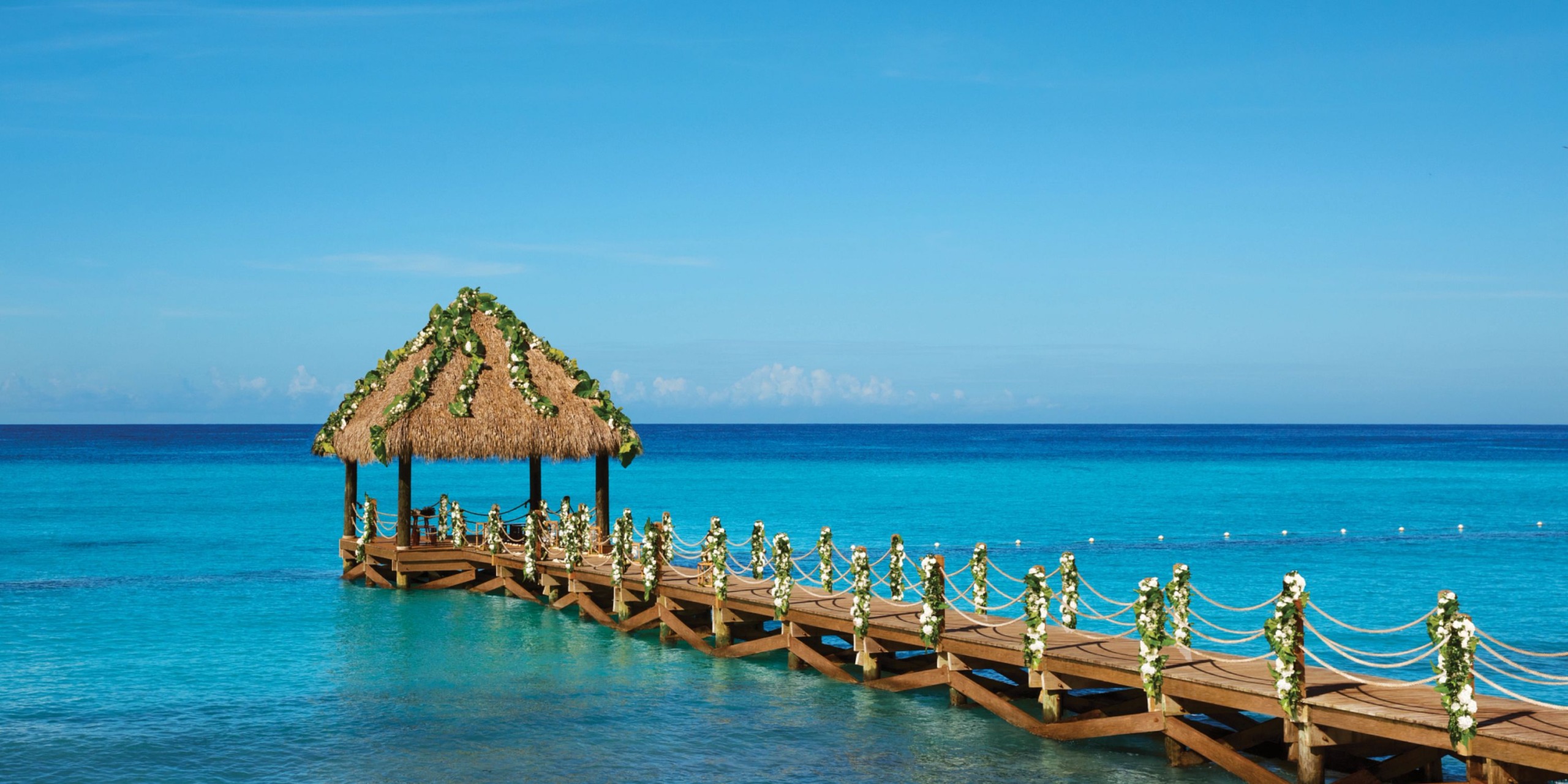 A wooden pier with a thatched-roof gazebo extends over the clear blue water under a bright sky at Dreams Dominicus.
