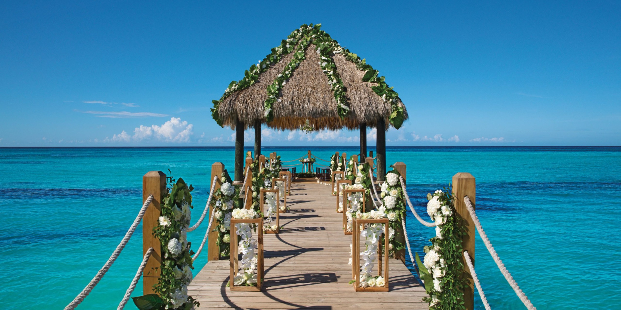 A wooden pier adorned with flowers extends to the thatched gazebo at Dreams Dominicus, set above clear blue waters beneath a brilliant sky.
