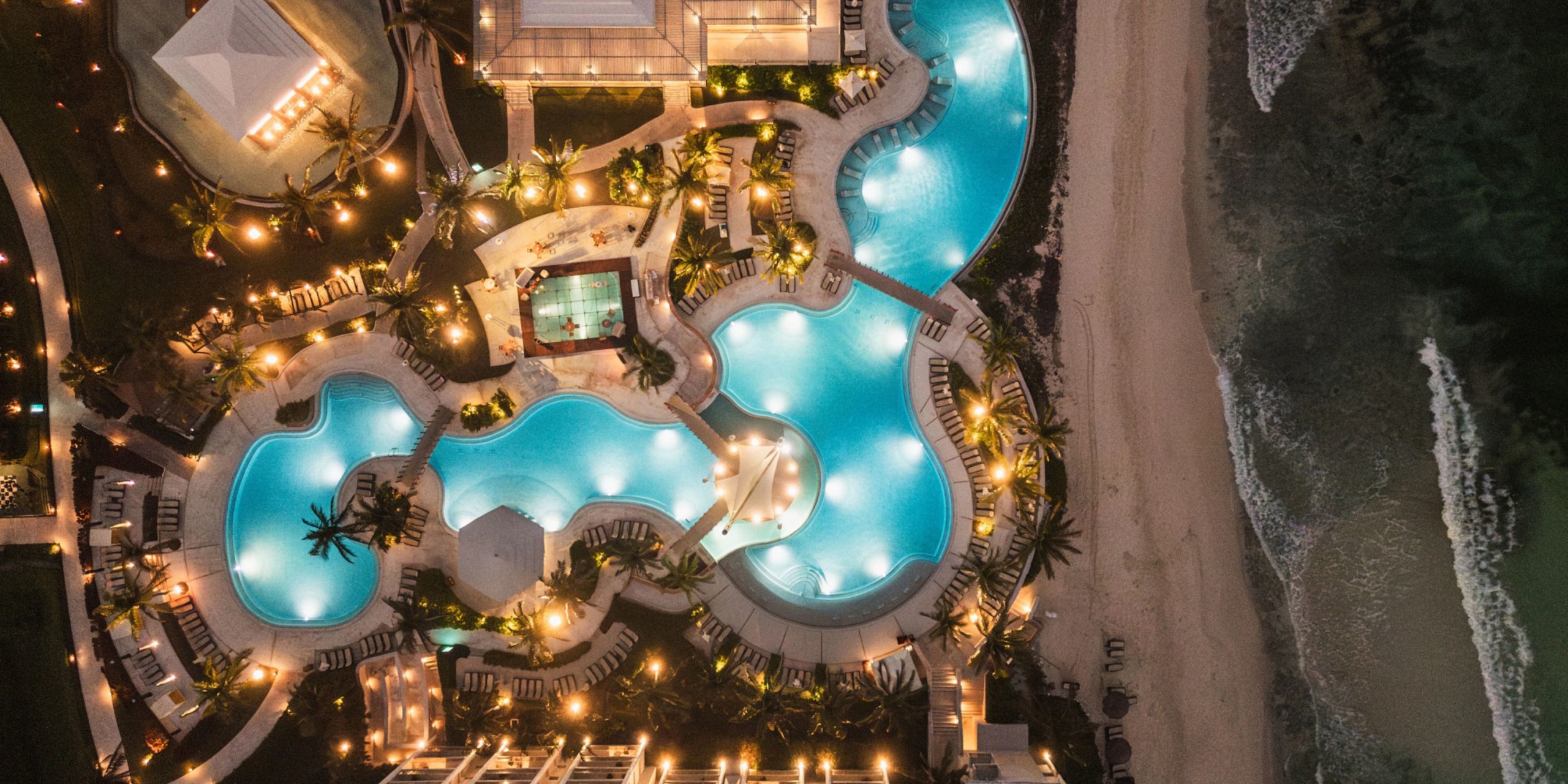 Aerial view of the illuminated resort pools at night at Dreams Jade Riviera Cancun, with a sandy beach and ocean waves on the right side. Palm trees and walkways surround the pools.