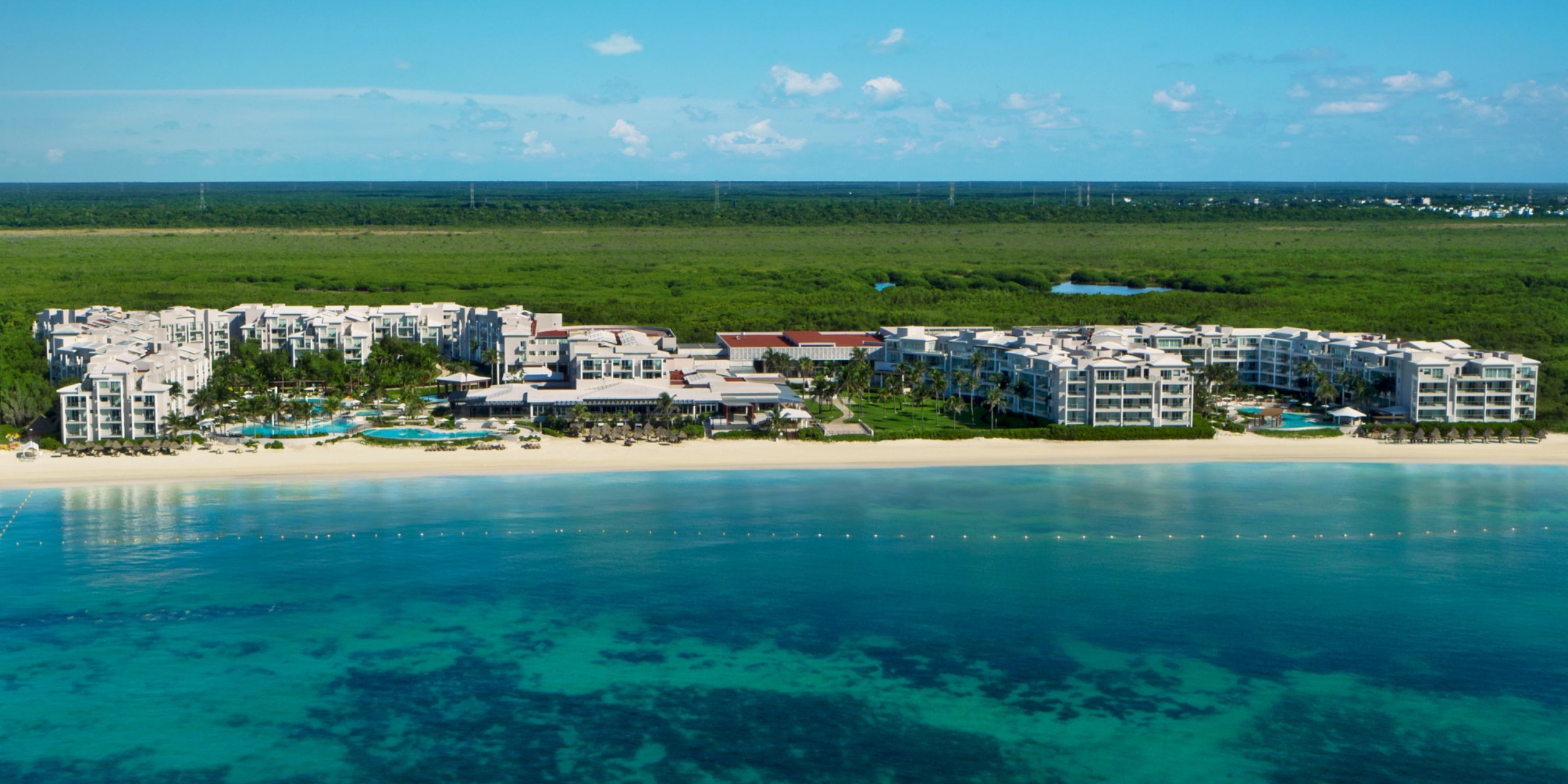 Aerial view of Dreams Jade Riviera Cancun, a beachfront resort featuring multiple buildings and pools adjacent to a white sandy beach and the blue ocean, with lush green vegetation visible in the background.