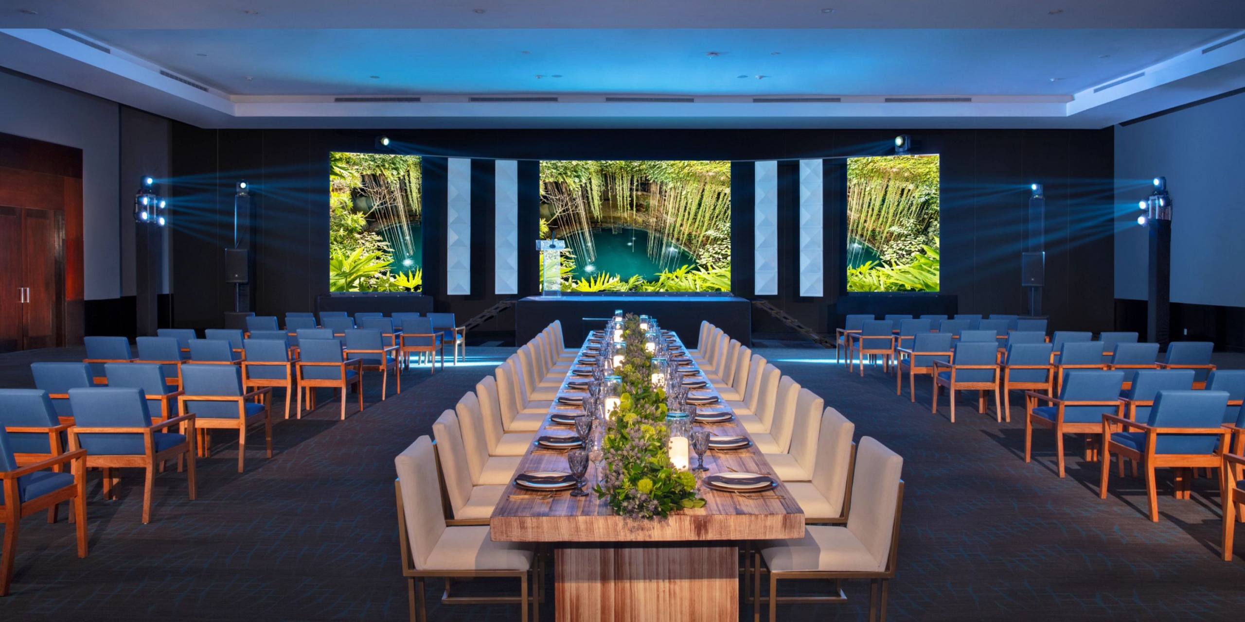 The Dreams Jade Riviera Cancun conference room features a long dining table surrounded by chairs, with large screens showcasing a tranquil nature scene of trees and water. Additional rows of seating are arranged to face the screens.
