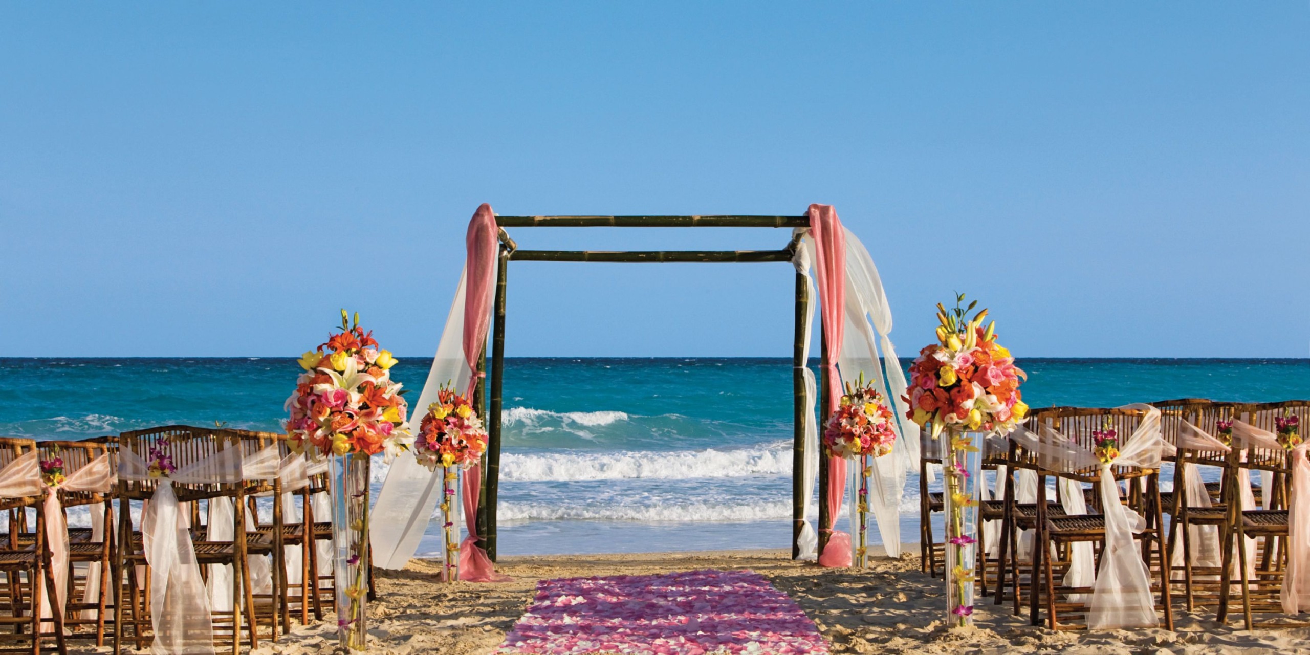 The Dreams Jade Riviera Cancun offers a picturesque beach wedding setup featuring a wooden arch adorned with flowing fabric, exquisite floral arrangements, and rows of chairs lining a pink petal-strewn aisle that leads to the breathtaking ocean view.