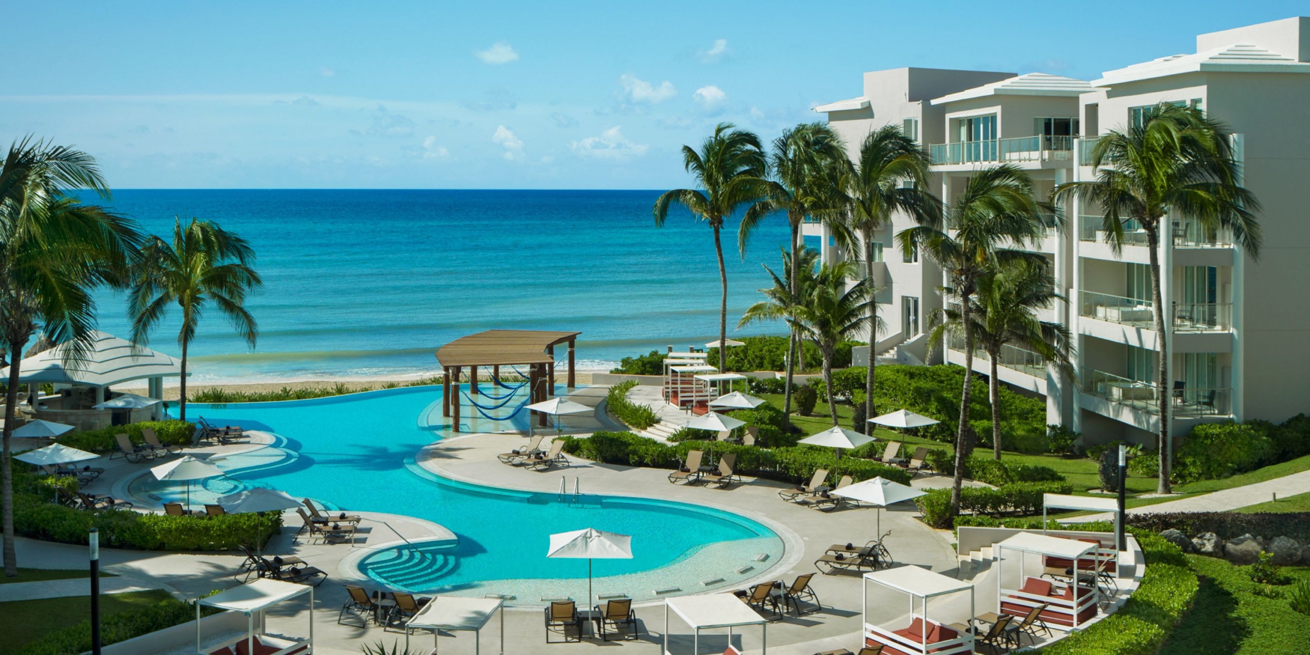 The Dreams Jade Riviera Cancun features a curvy pool, surrounded by palm trees and sun loungers, offering an ocean view under a clear blue sky.