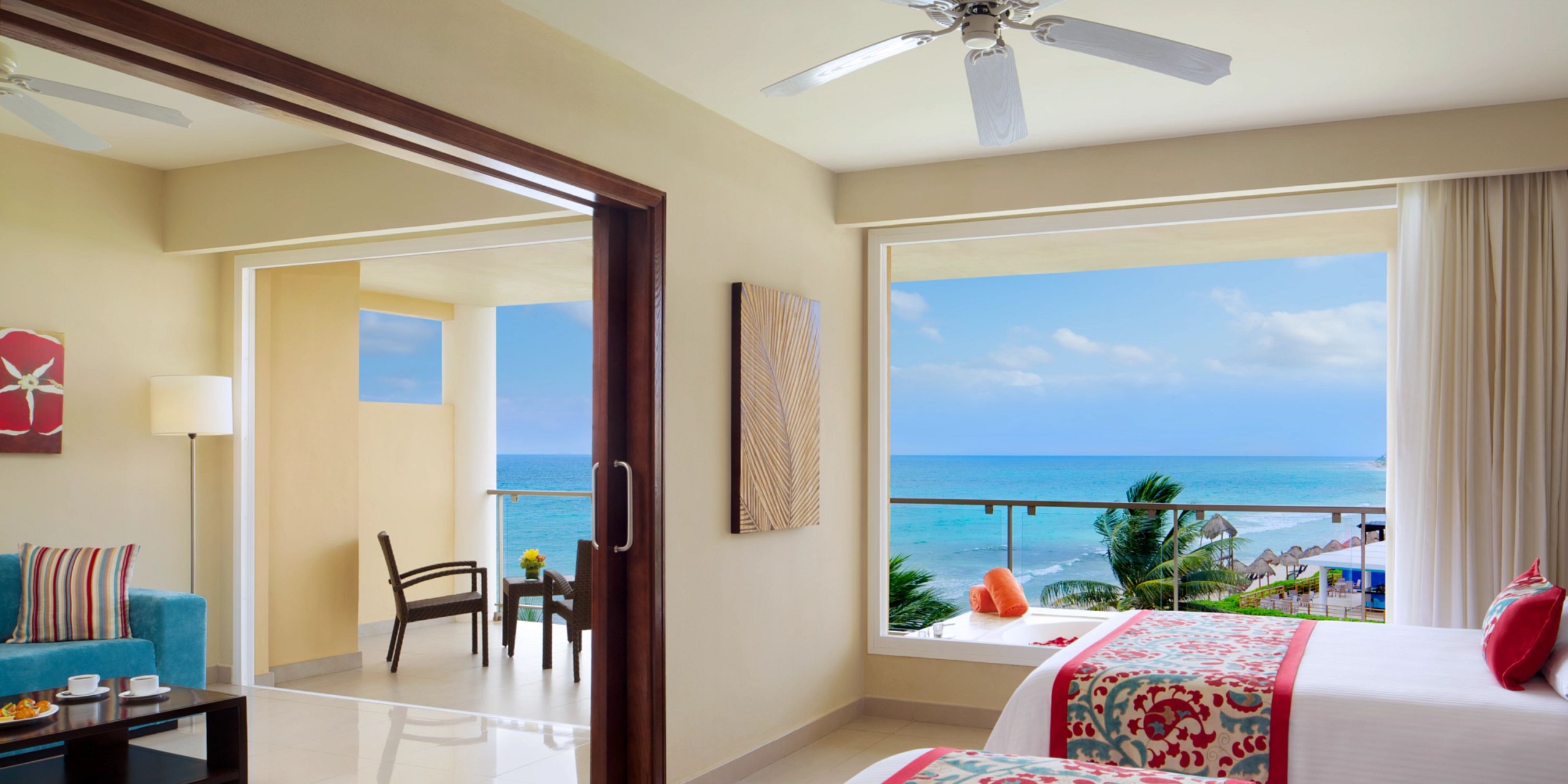 Experience a stay at Dreams Jade Riviera Cancun in a hotel room featuring two beds adorned with decorative pillows and offering a partial ocean view. The sliding door opens to a balcony equipped with chairs and a table, while a ceiling fan gently circulates the air above.